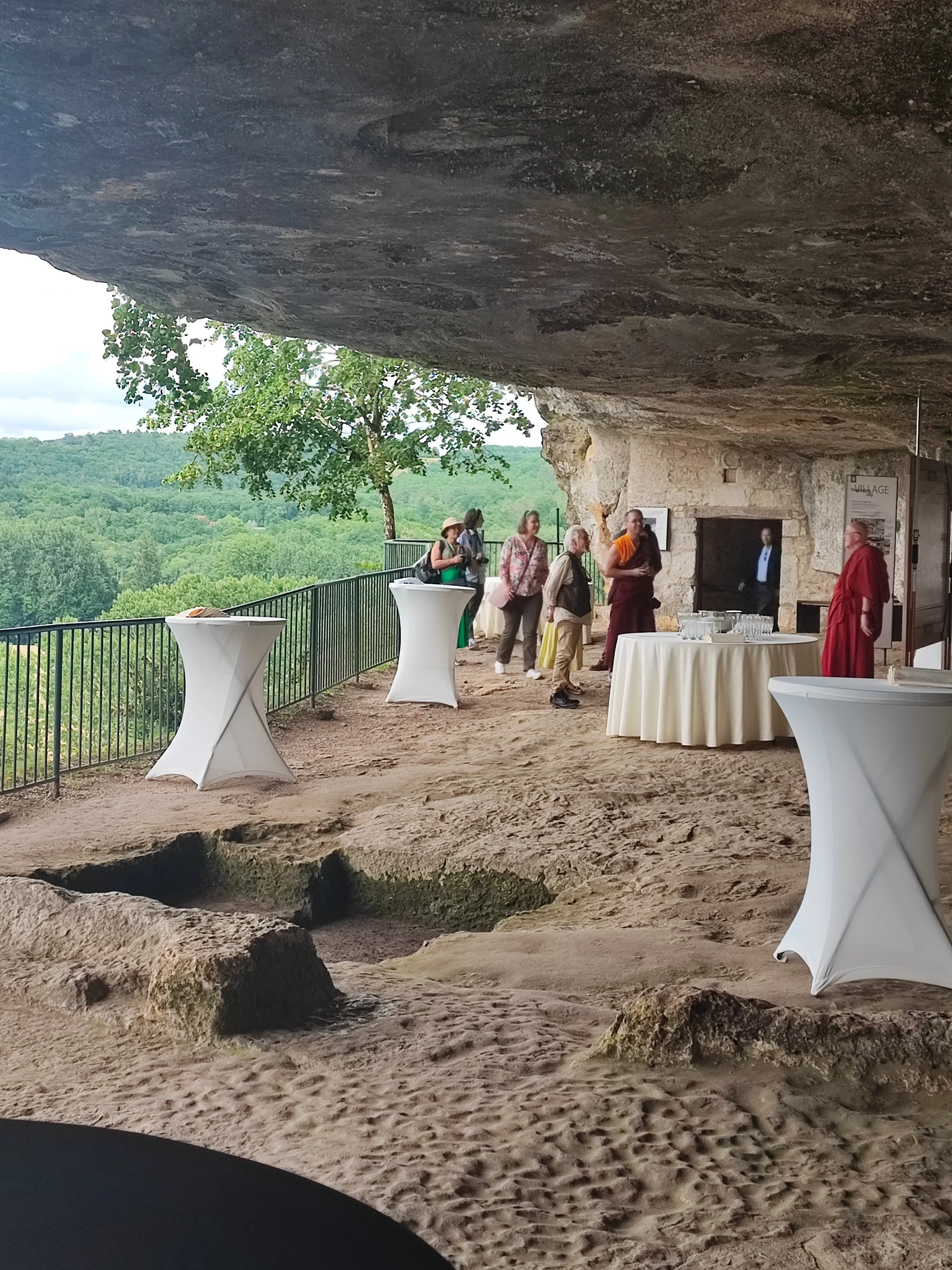 Un groupe de personnes se tient dans une structure construite à l'intérieur d'une grotte ou d'un site rocheux, avec une vue panoramique de la nature en arrière-plan. Il y a des tables hautes avec des nappes blanches, un écran d'information et une fig