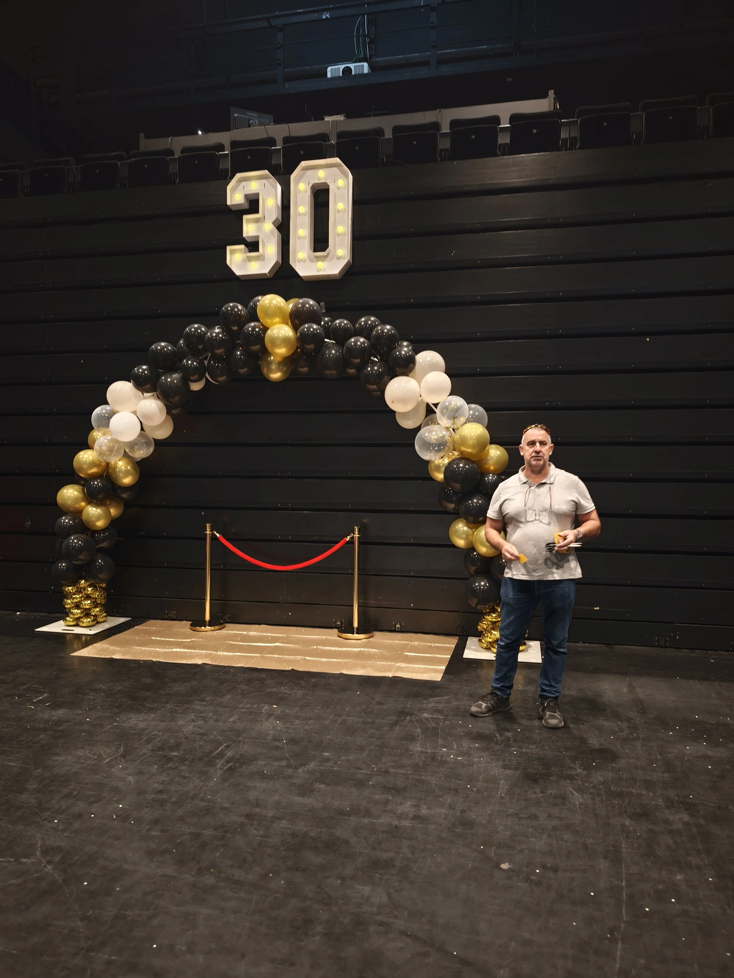 Décoration pour un anniversaire de 30 ans avec un ballon en forme d'arc noir, blanc, doré et transparent, un chiffre 30 lumineux fixé au mur noir, un tapis doré, des barrières rouges et un homme debout à côté de la décoration, dans une salle avec des