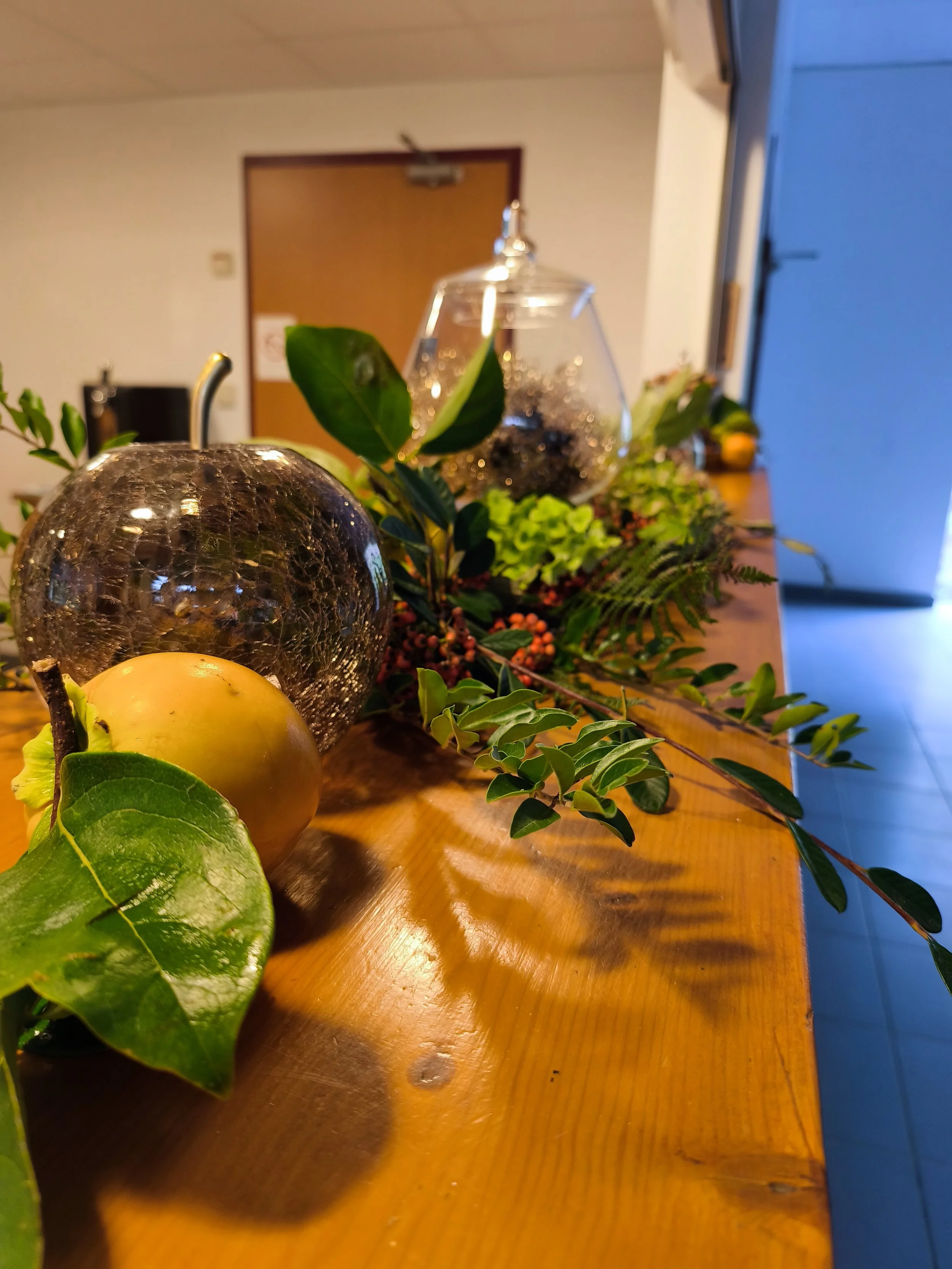 Décoration florale avec feuillage, boules en verre, un fruit jaune, et un vase en verre sur une table en bois.