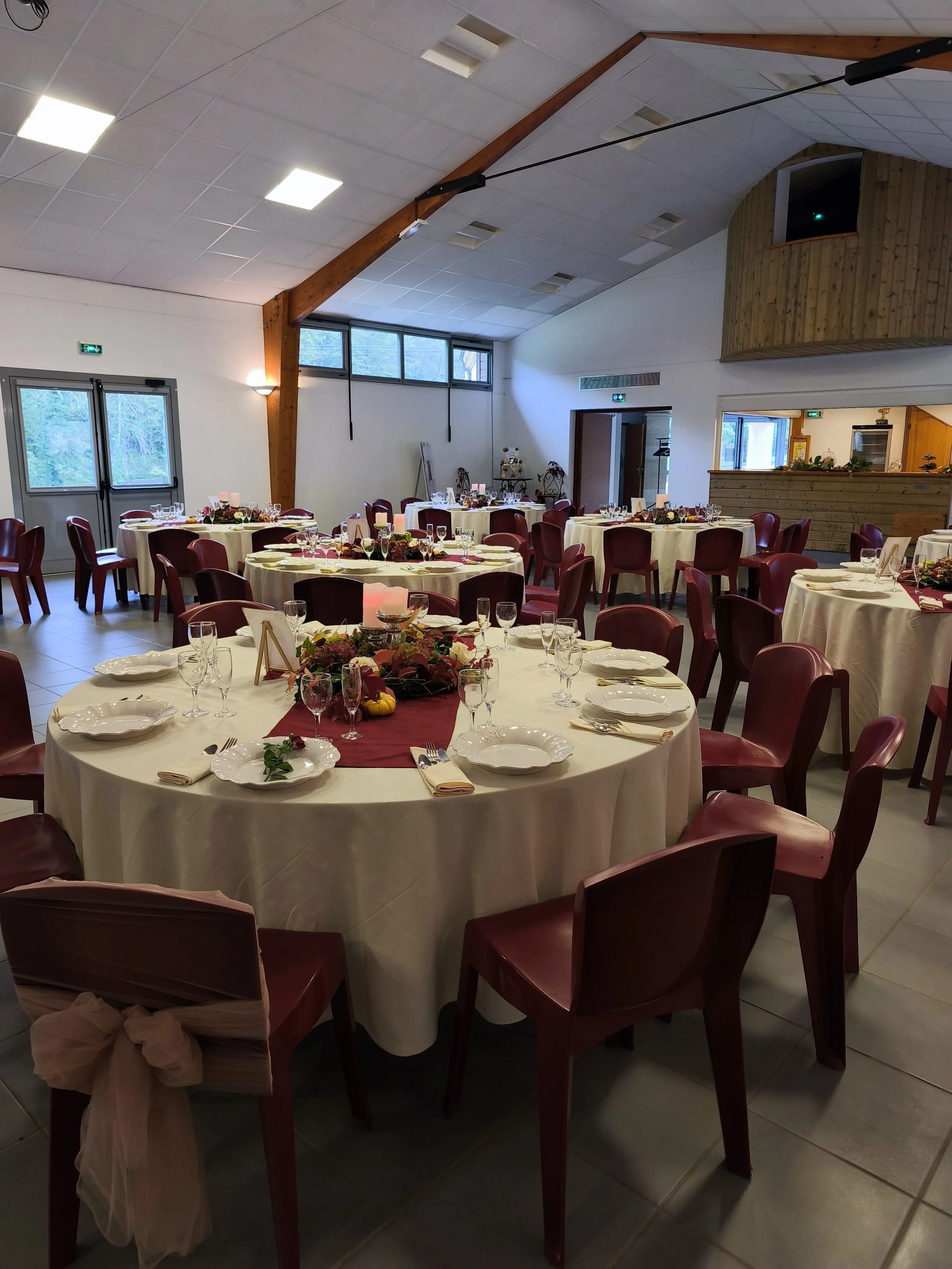 Salle de réception décorée pour un événement avec tables rondes, nappes blanches, vaisselle, verres et centres de table fleuris, avec des murs en bois et des fenêtres.
