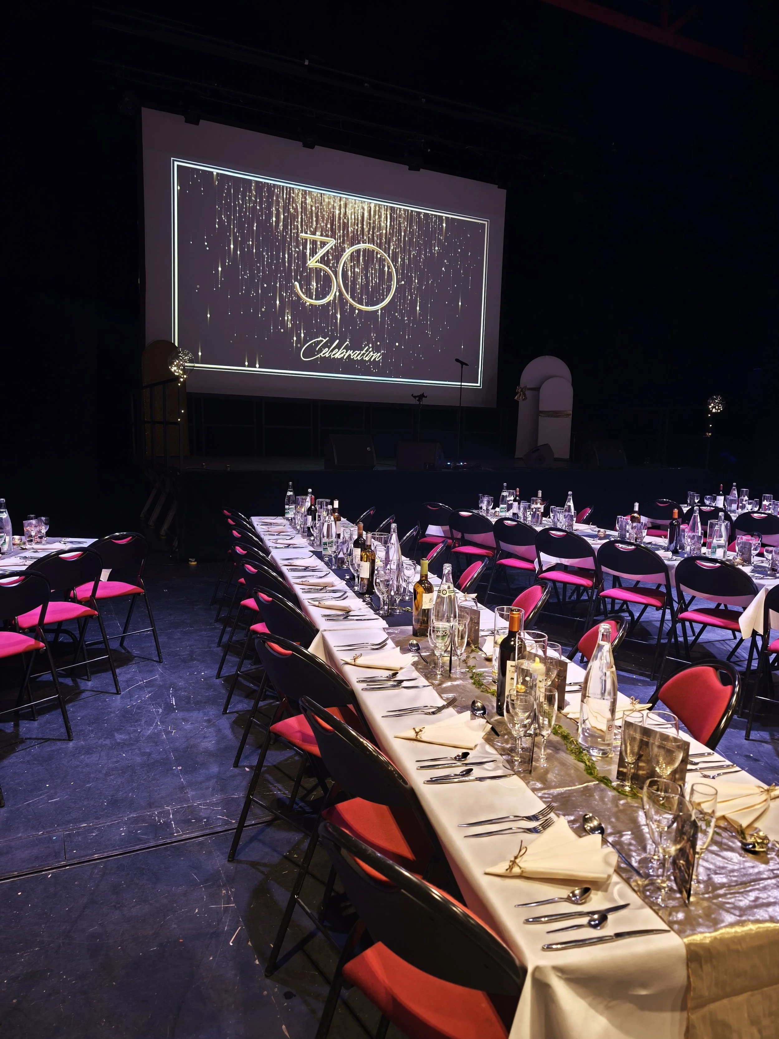 Salle décorée pour une célébration du 30e anniversaire avec une grande table, bouteilles de vin et verres, un grand écran affichant "30th Celebration" avec des feux d'artifice dorés, en arrière-plan des lumières décoratives.