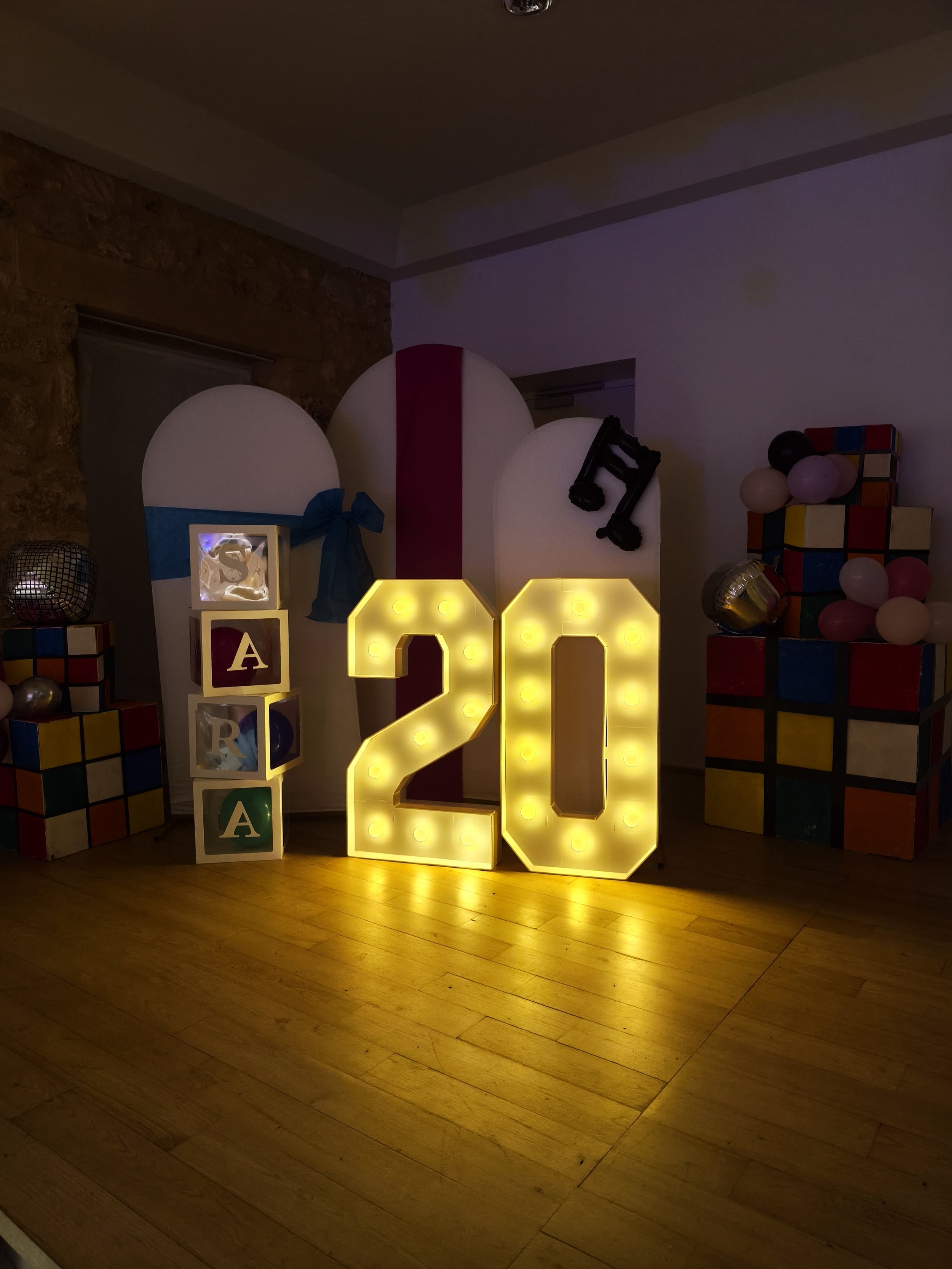 Décoration lumineuse de anniversaires avec le chiffre 20 en lettres colorées et en chiffres lumineux, entourée de ballons roses, noirs, blancs, et cubes colorés.