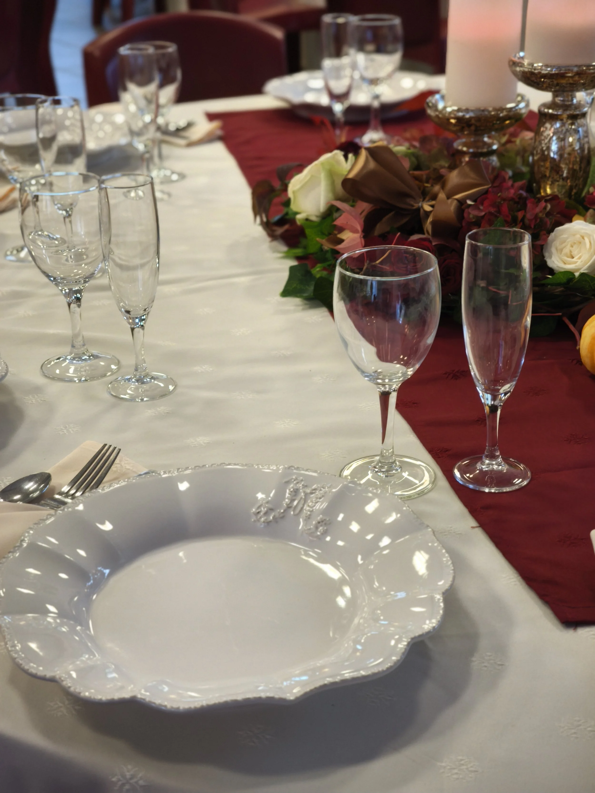 Une table de dîner élégante avec plusieurs verres à vin, des assiettes en porcelaine blanche avec un motif délicat, une nappe blanche et une décoration florale avec des roses blanches et des fleurs rouges. Des bougies ornées de pièces en or ajoutent 