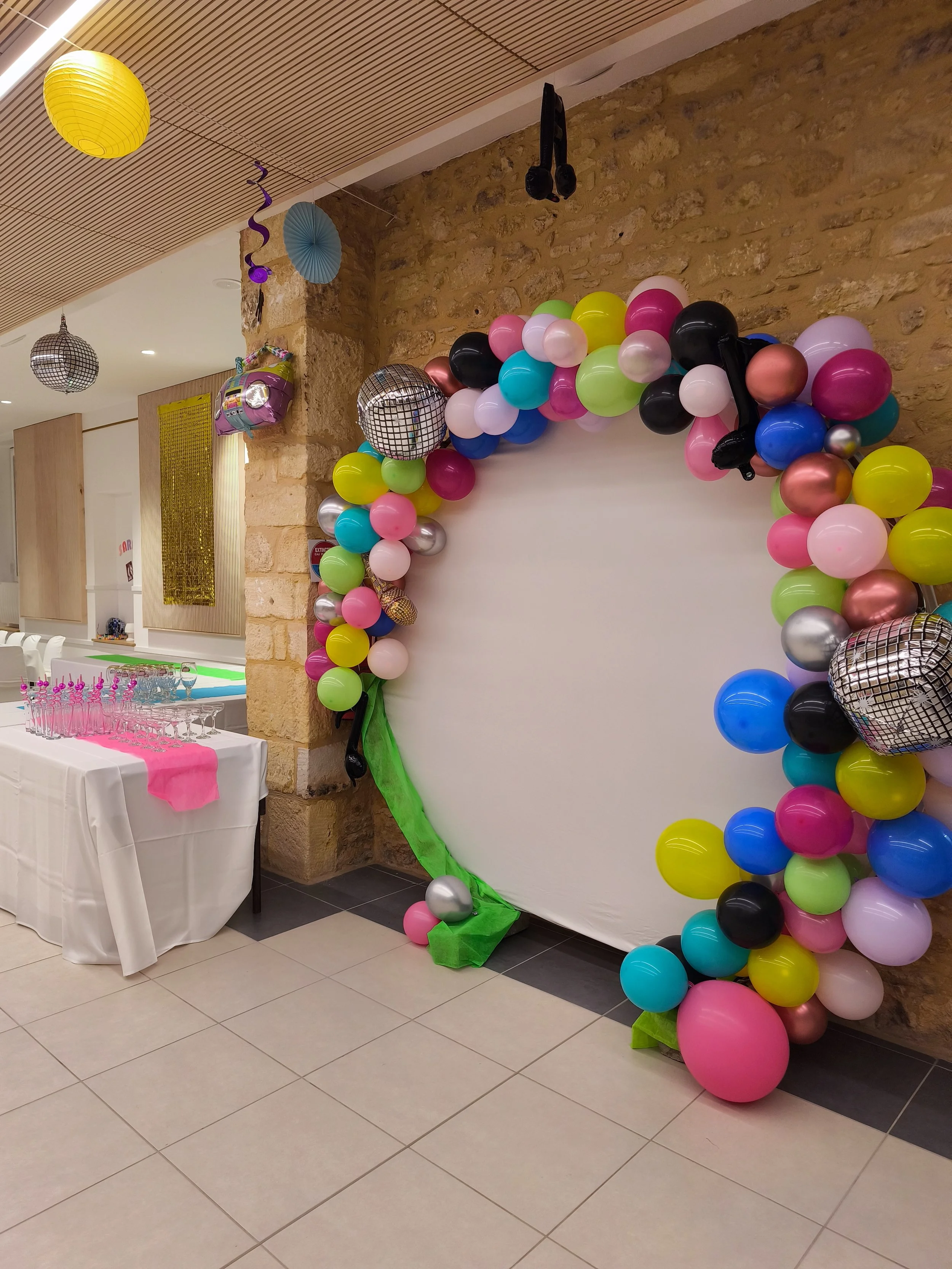 Décoration de fête avec un ballonnet multicolore en arc, plusieurs ballons de différentes couleurs, deux ballons disco en miroir, et un fond blanc. La décoration est fixée à un mur en pierre dans un espace intérieur avec un plafond en bois.