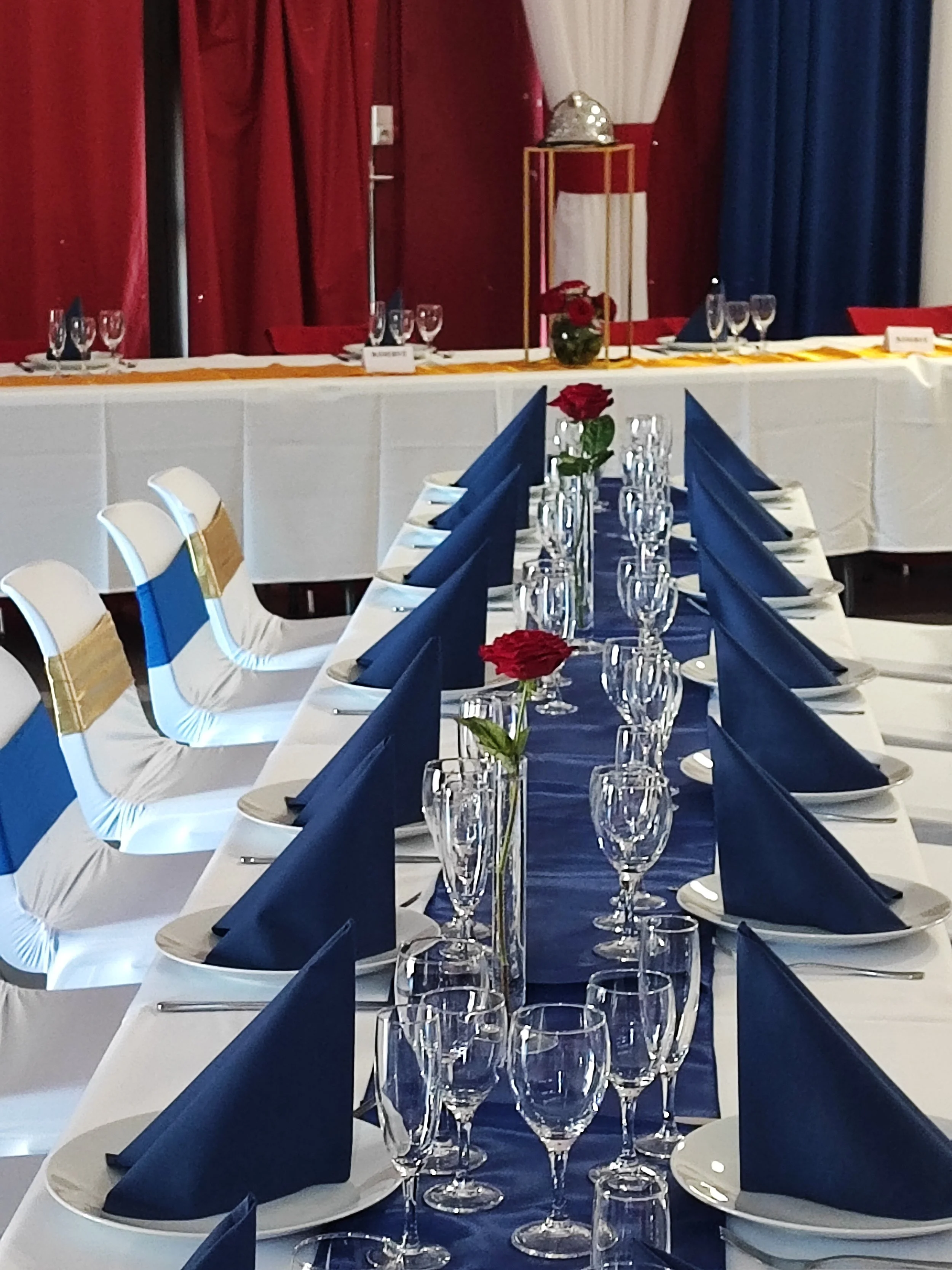 Une salle décorée pour un événement avec une longue table dressée, nappes blanches, serviettes bleues en forme de triangle, verres à vin et roses rouges en vase, en arrière-plan un stand avec un plateau en argent et un vase avec des roses rouges.