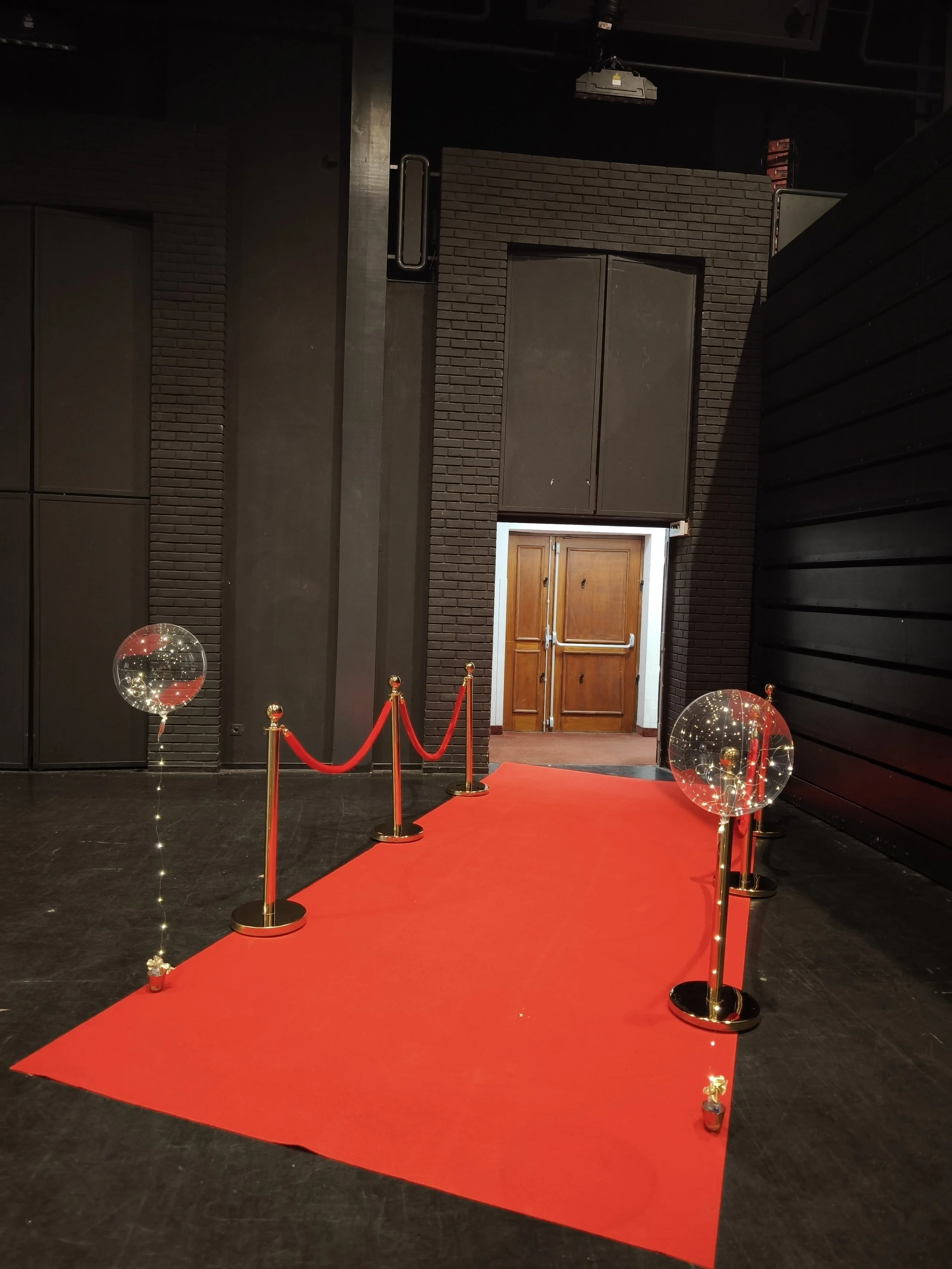 Un tapis rouge avec des barricades dorées et des ballons transparents décorés de guirlandes lumineuses, dans un espace intérieur avec des murs sombres, prêt pour une cérémonie ou un événement spécial.