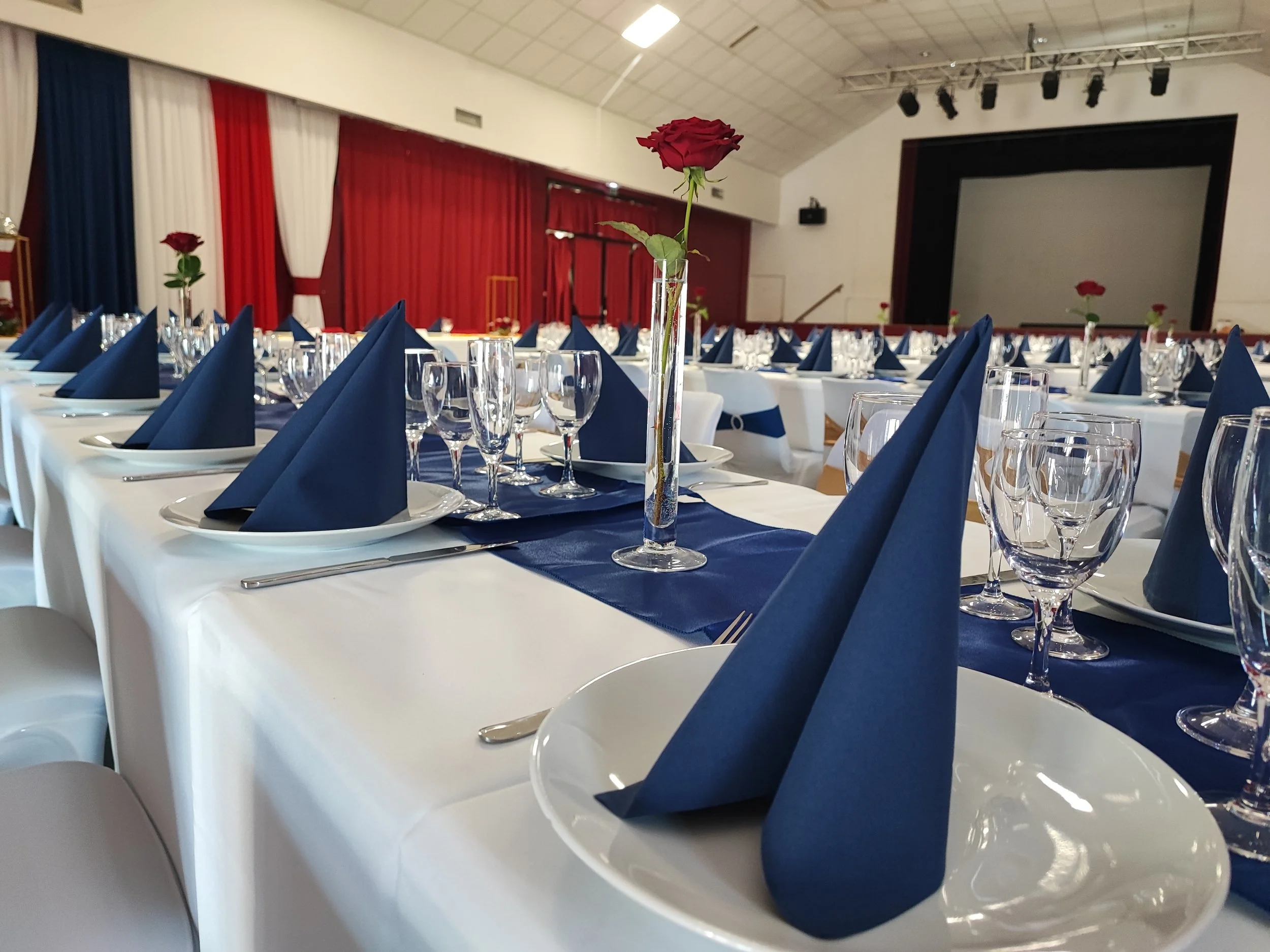Salle de banquet décorée pour un événement, avec table ronde, nappes blanches, serviettes bleues pliées, verres à vin, flûtes à champagne, et violettes. Il y a un centre de table avec une rose rouge dans un vase transparent. Murs avec rideaux rouges,