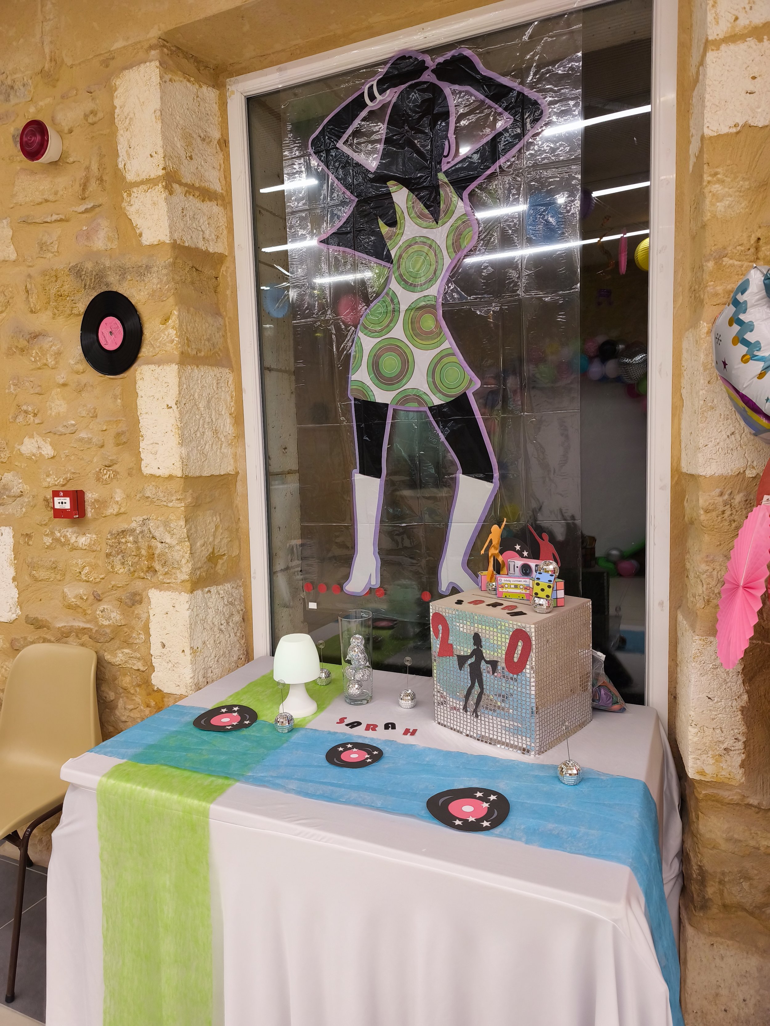 Décoration de fête avec un mur en pierre, une table couverte d'une nappe blanche avec une bande verte, une lampe blanche, des disques en carton à disque de vinyle, un cadeau avec un thème de boîte miroir, un ballon rose, une partie de décorating avec