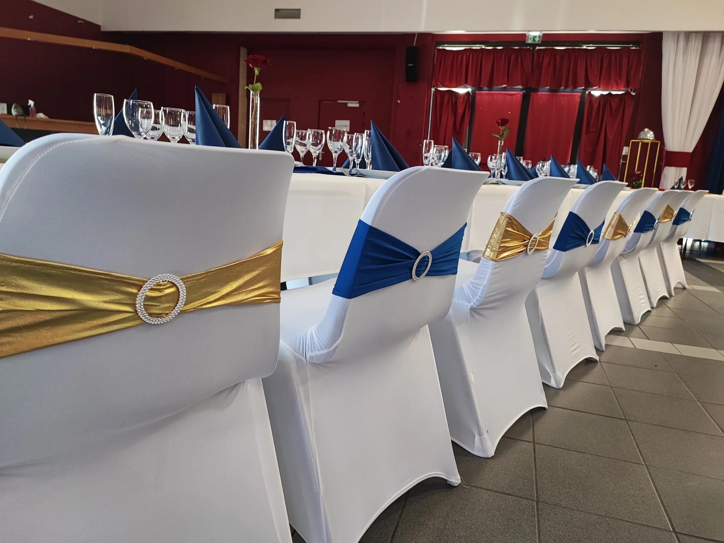 Salle décorée avec des tables de banquet préparées pour un évènement, avec des verres, des serviettes bleues, et des cheminements dorés avec des broches de bijoux, avec des chaises blanches et des décorations florales rouges.