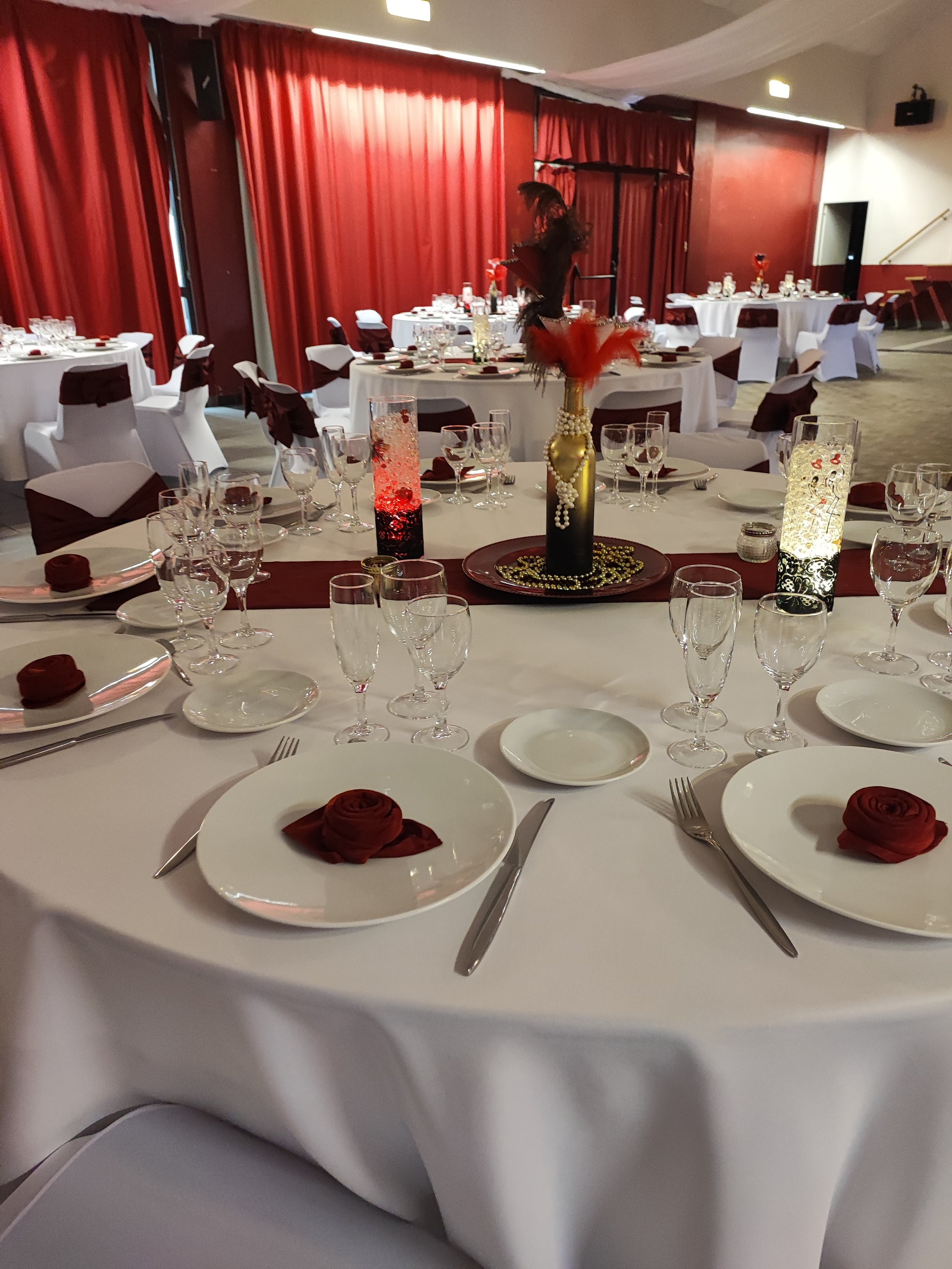 Salle de banquet élégante avec plusieurs tables rondes décorées de nappes blanches, de centres de table avec des vases noirs contenant des plumes rouges et ornementées de perles. Les chaises sont habillées de housses blanches avec des rubans rouges. 