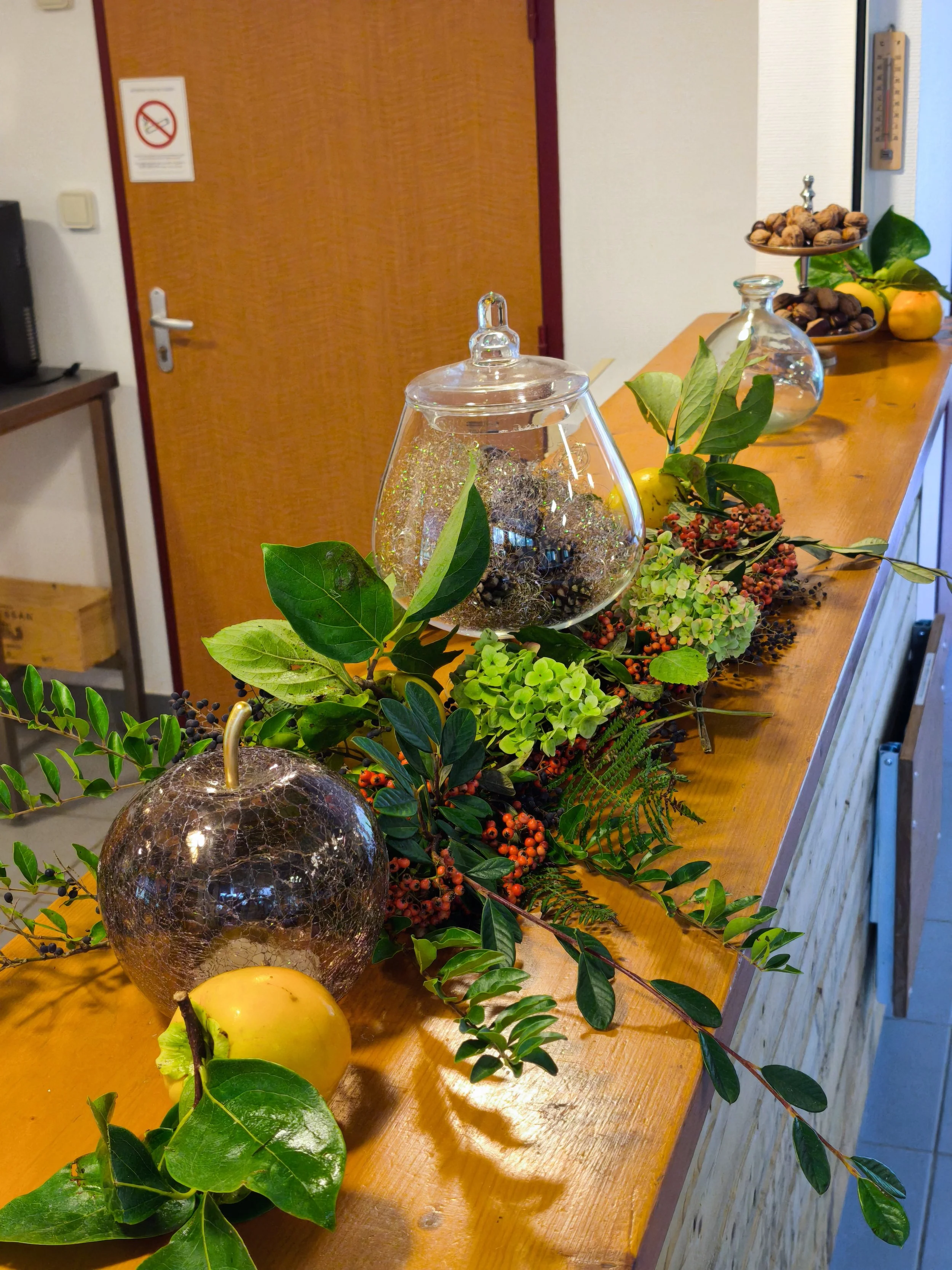 Décoration de table avec des plantes vertes, des fruits jaunes, et des contenants en verre, avec un arrière-plan d’une porte en bois.