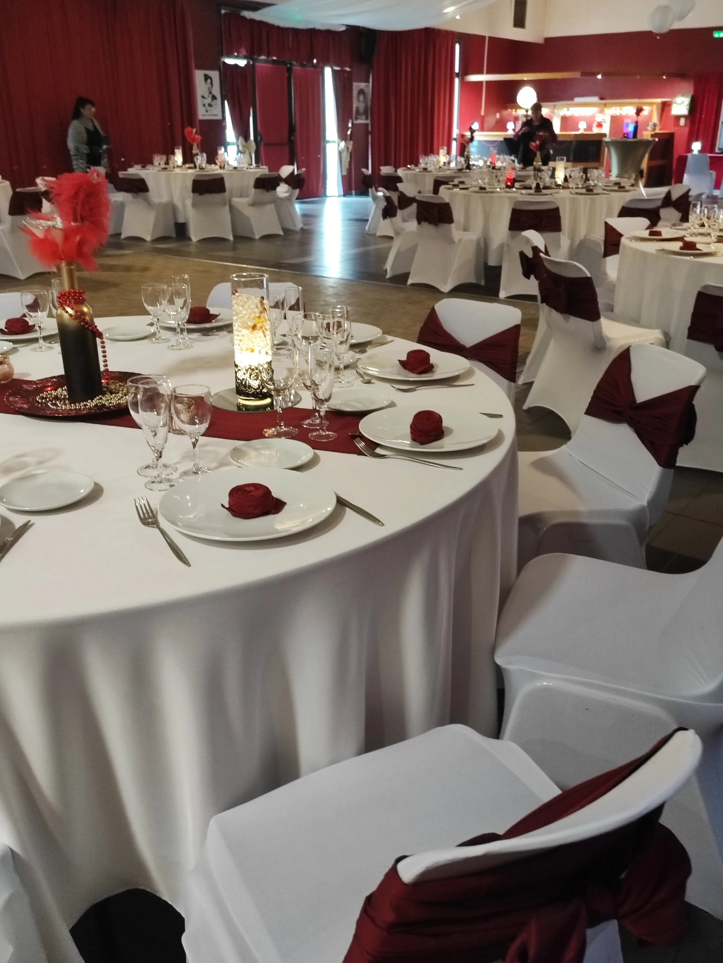 Salle de réception décorée pour un événement, avec des tables rondes ornées de centres de table rouges et de vaisselle élégante. Chaises blanches avec rubans bordeaux. Un grand espace avec des rideaux rouges foncés et un éclairage chaleureux.