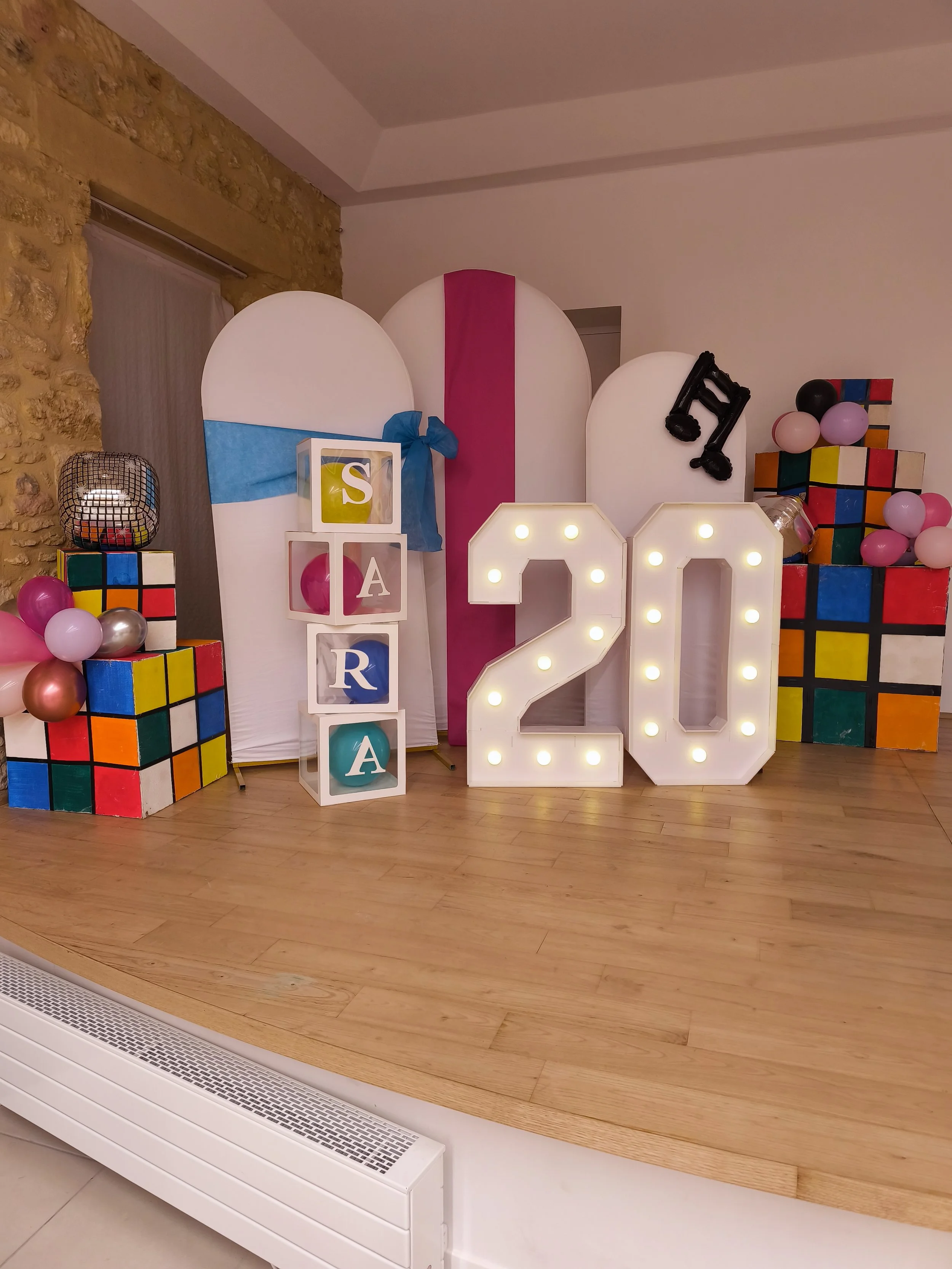 Décor de fête avec des ballons roses, noirs, et métalliques, des blocs colorés, des lettres éclairées formant le chiffre 20, et des boîtes avec des lettres 'S', 'A', 'R', 'A'.