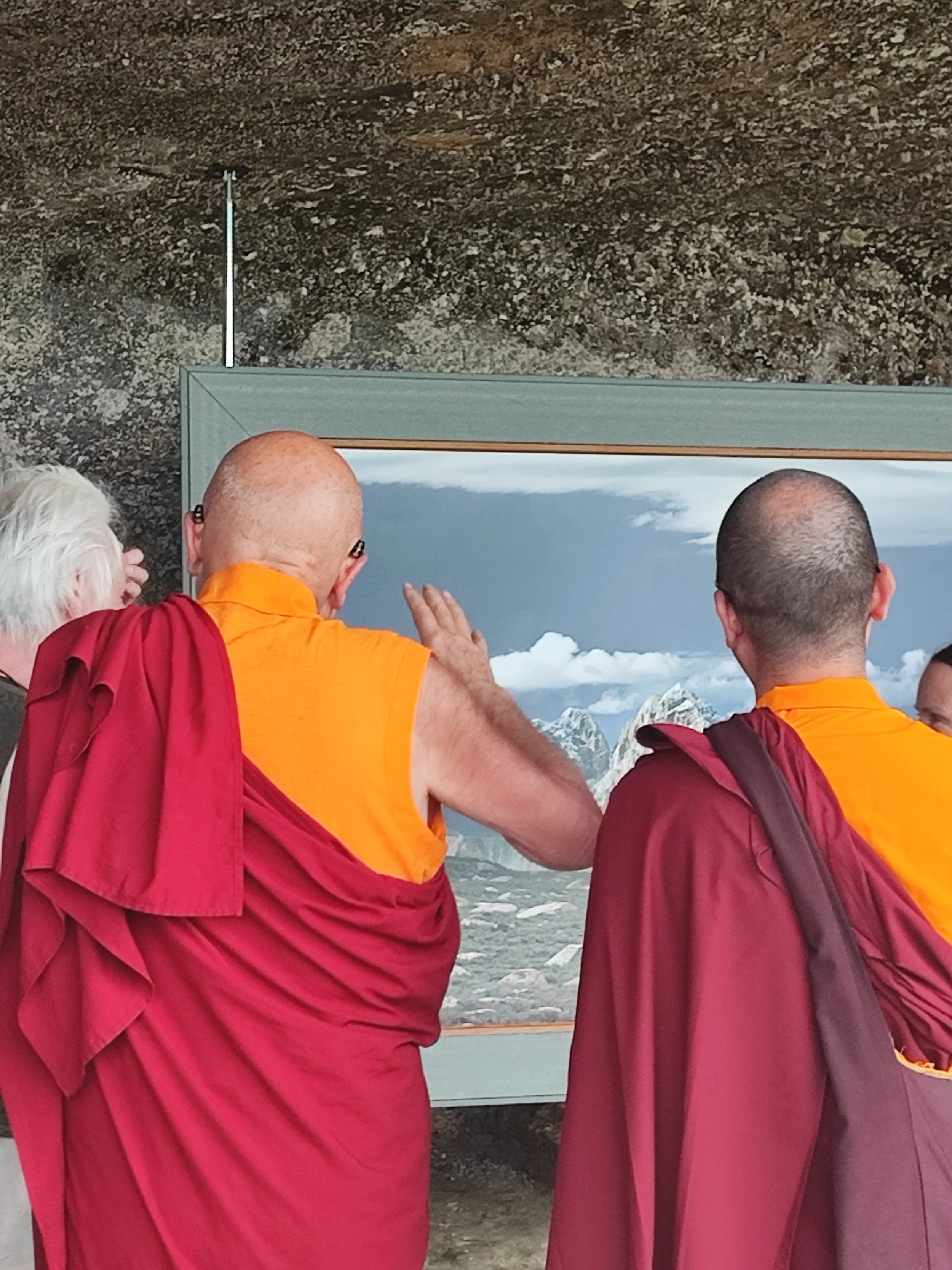 Trois personnes portent des habits de moines tibétains de couleur rouge et orange, regardent une photographie de montagnes enneigées.
