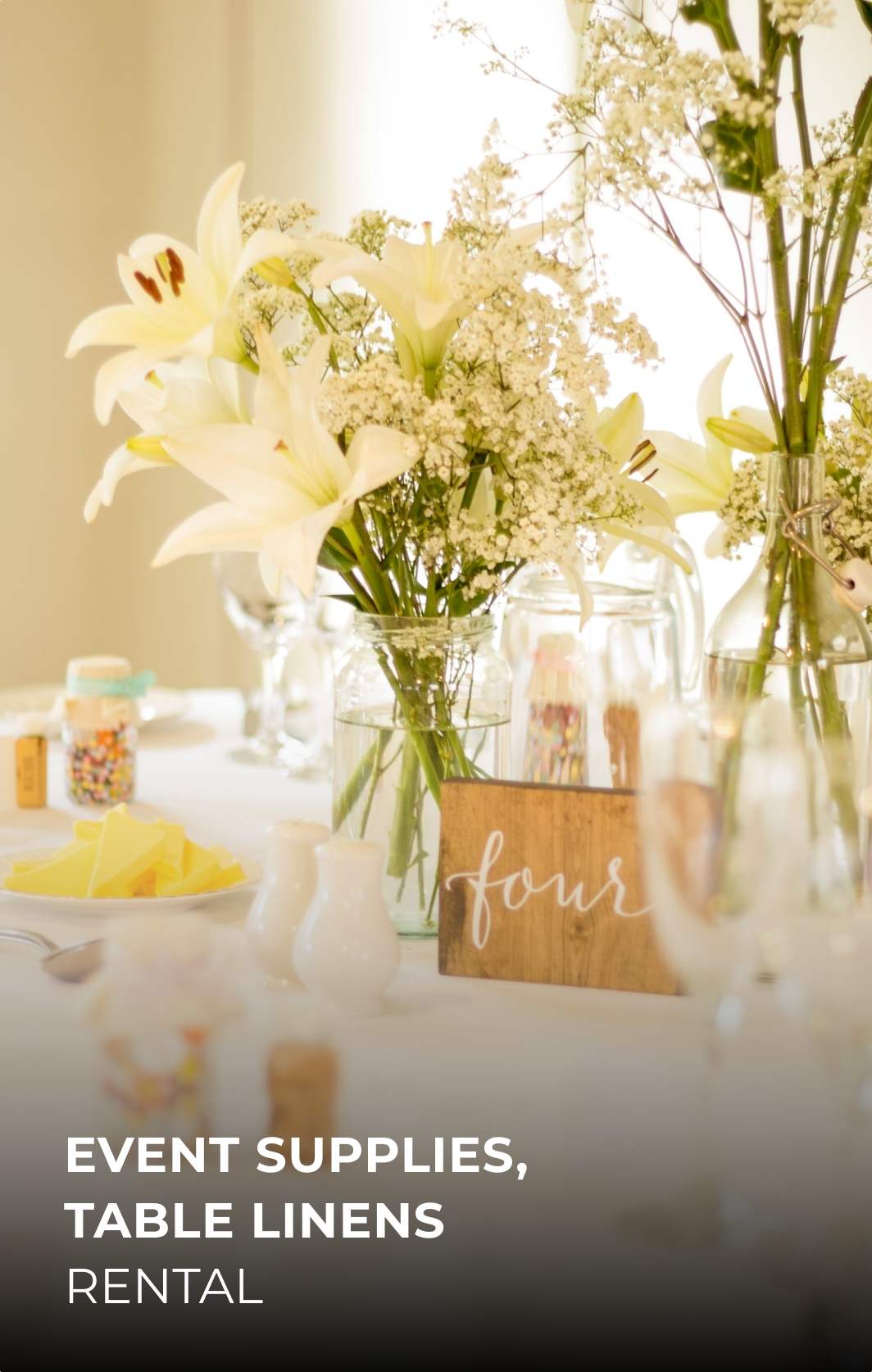 Vase en verre avec des fleurs blanches, notamment des lys et des gypsophiles, décorant une table d'événement avec des contenants en verre et des petits objets colorés.