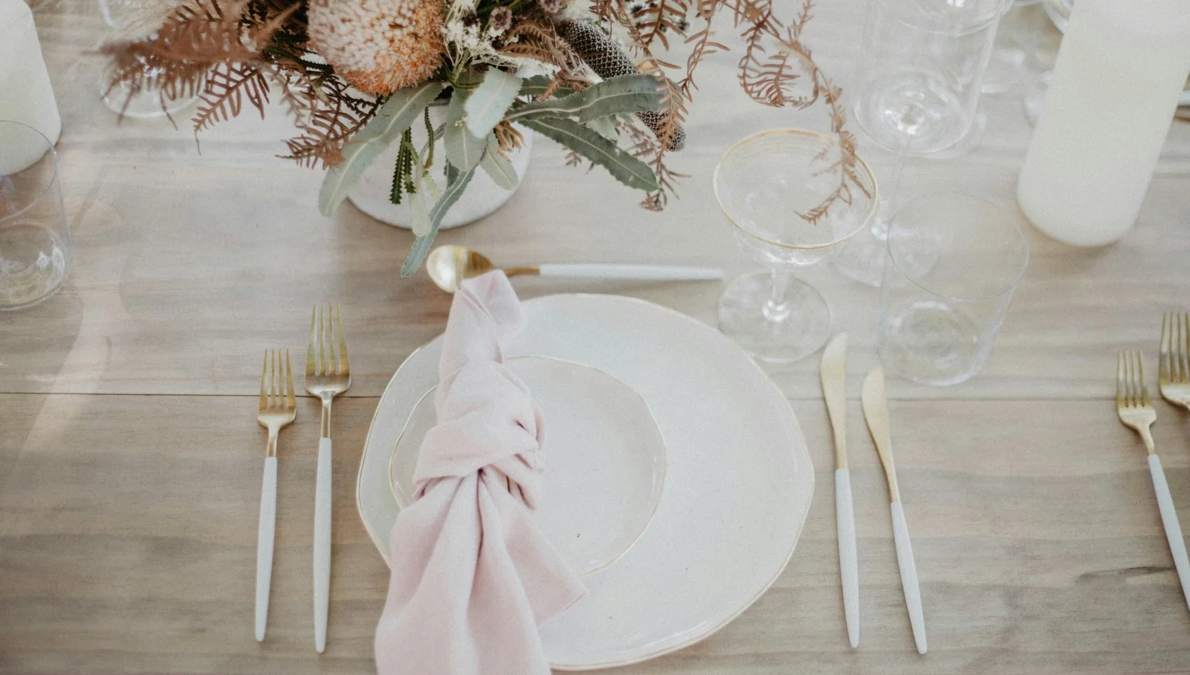 Table élégamment décorée avec une nappe en bois clair, un centre de table floral avec des fleurs et feuillages dans un vase blanc, des assiettes blanches avec une serviette rose enroulée, des fourchettes à gauche, des couteaux et cuillères à droite, plusieurs verres à vin et un verre d'eau, et des bougies blanches en arrière-plan.