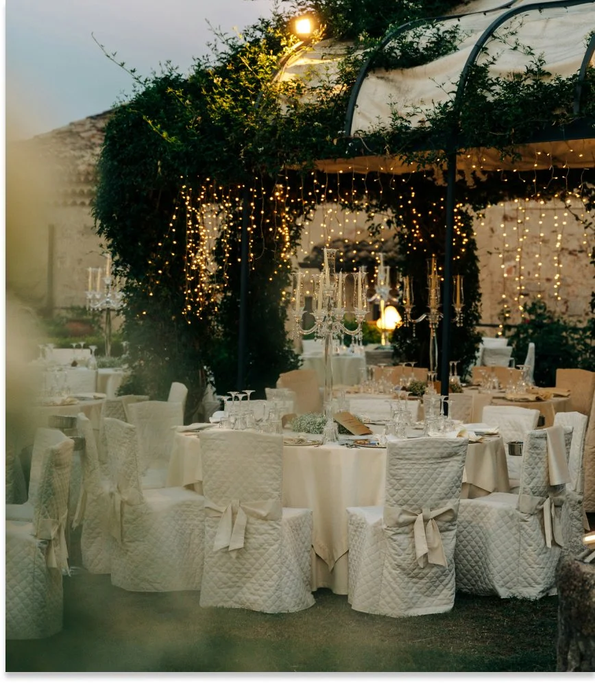 Un lieu de réception en plein air avec des tables rondes dressées avec de la vaisselle et des chandeliers. Des guirlandes lumineuses décorent la structure et la végétation environnante, créant une ambiance festive et élégante à la tombée de la nuit.