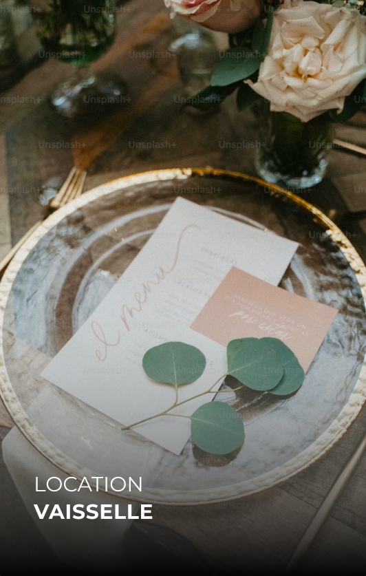 Une assiette en verre sur une table en bois avec un menu, un bout de carte et une branche de feuilles vertes sur un plate serviette blanc écru, avec un vase de fleurs en arrière-plan.