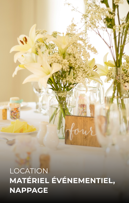 Vasque avec un arrangement floral composé de lys blancs et d'autres fleurs blanches, sur une table décorée avec des éléments pour un événement, dans une lumière naturelle.