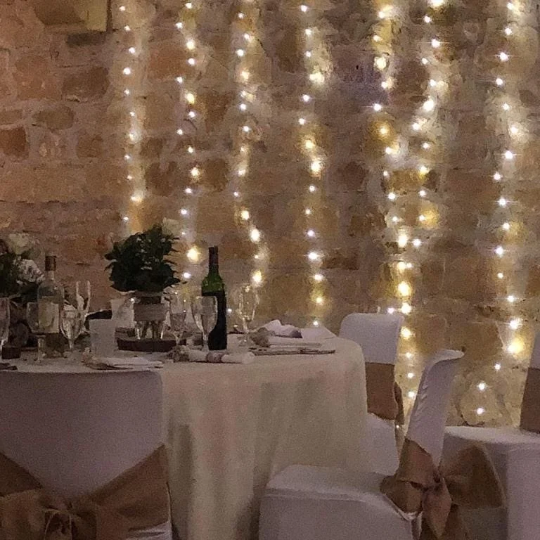 Une table décorée pour un repas, avec des verres, assiettes, flûtes à champagne, et une bouteille de vin ou champagne, entourée de chaises avec des nœuds en tissu. Derrière, des guirlandes lumineuses suspendues créent une ambiance chaleureuse.