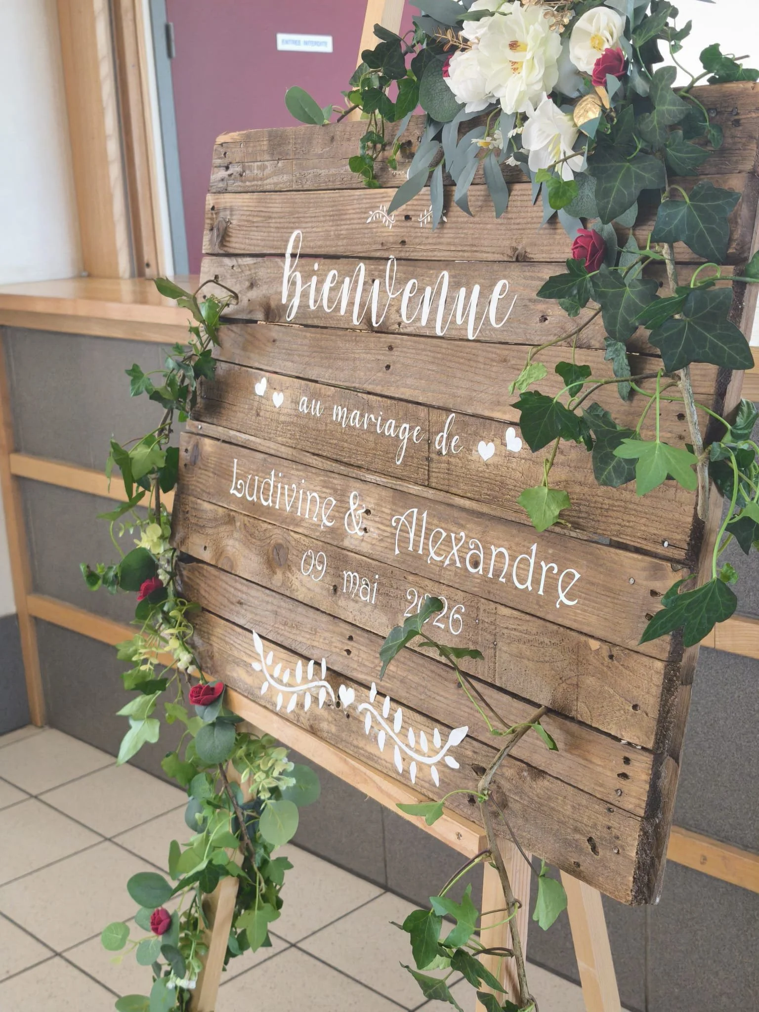 Panneau en bois avec le mot 'Bienvenue' écrit en blanc, décoré de fleurs blanches et rouges, ainsi que de feuilles vertes, en style rustic.