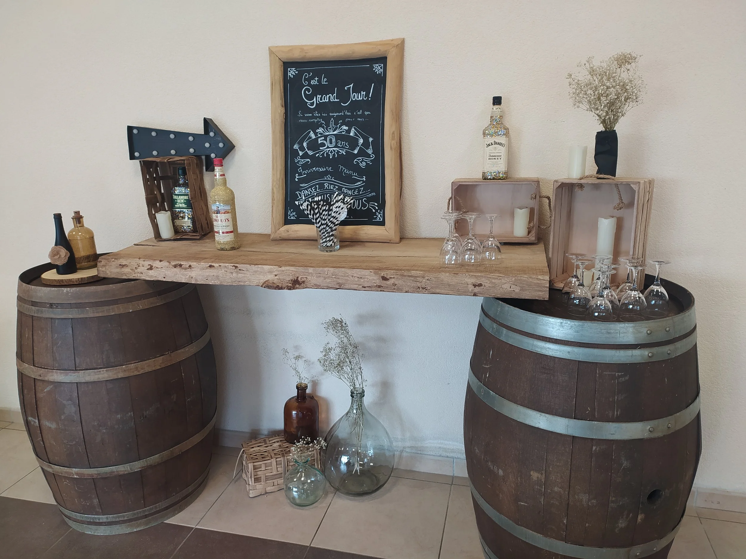 Décoration de fête avec deux tonneaux en bois servant de support pour un plateau de bouteilles d'alcool, une déco de journaux, des verres à vin, des bougies, des fleurs séchées et un tableau noir avec message écrit en français pour un anniversaire de 50 ans.