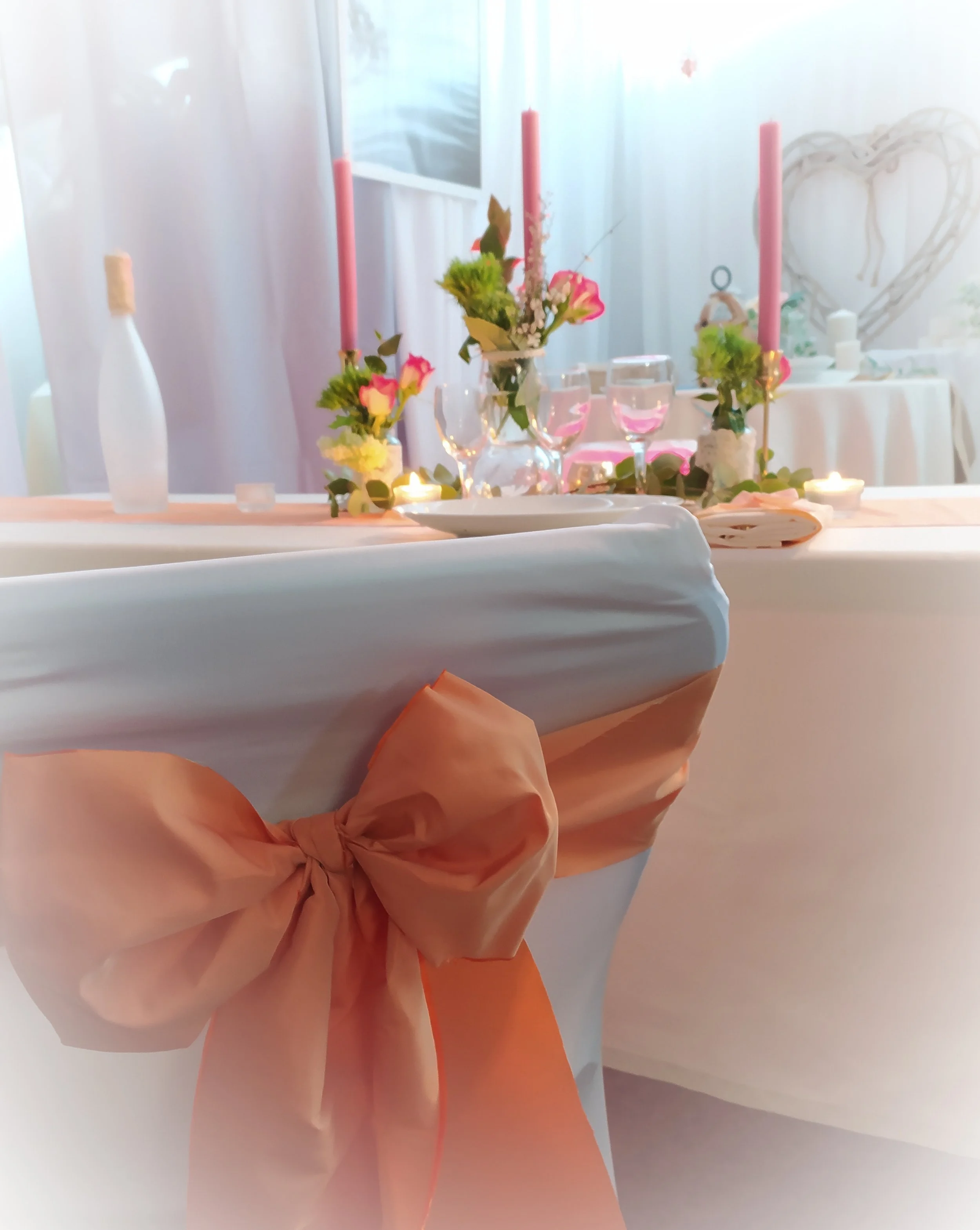 Table décorée pour une fête ou un événement avec des fleurs, des bougies, des verres à vin et des bougies roses, et des éléments de décoration blancs et roses.