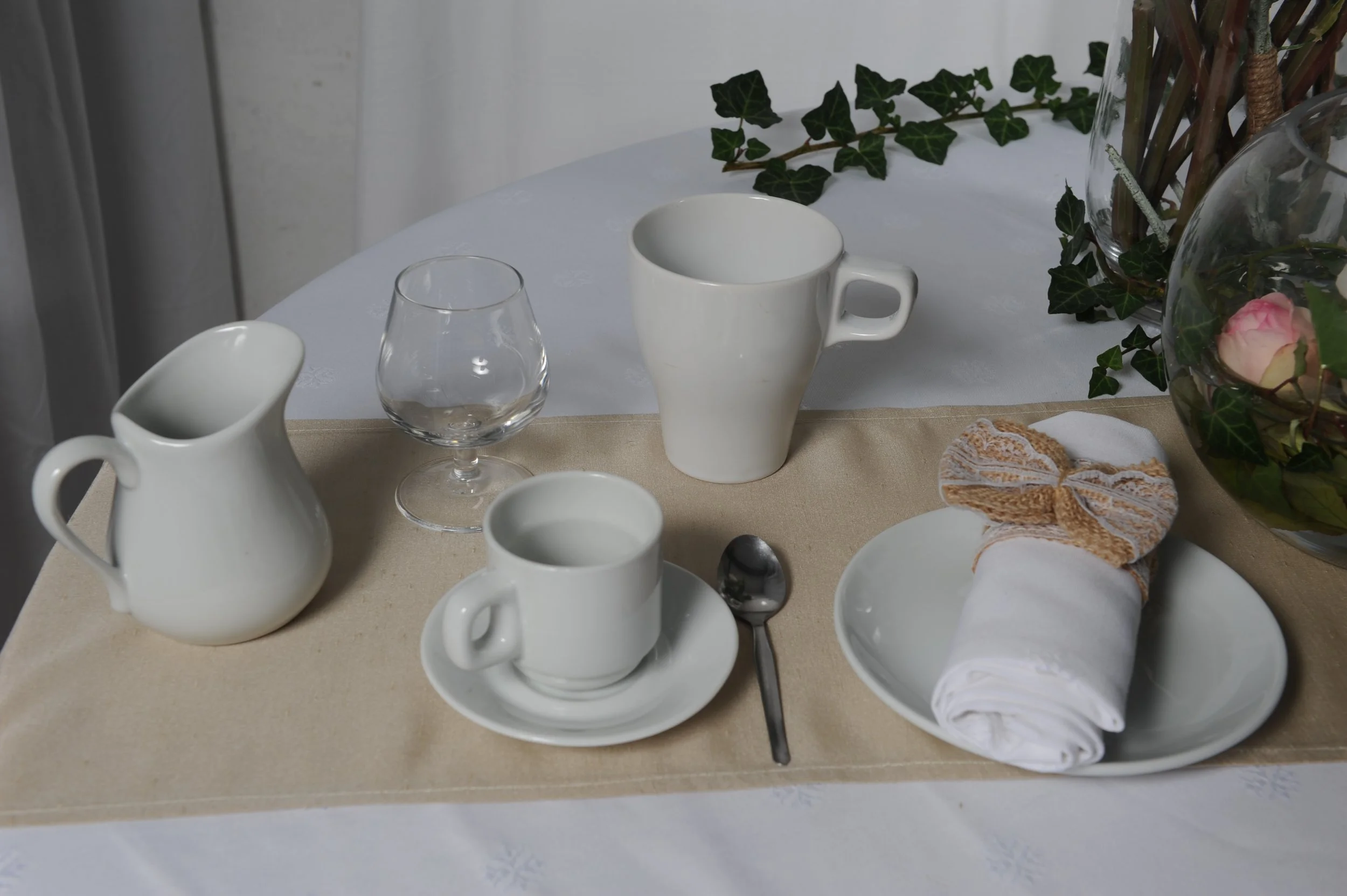 Service de vaisselle avec tasses, carafes, verre et cuillère, décoré avec des plantes vertes et une rose, sur une table avec une nappe beige.