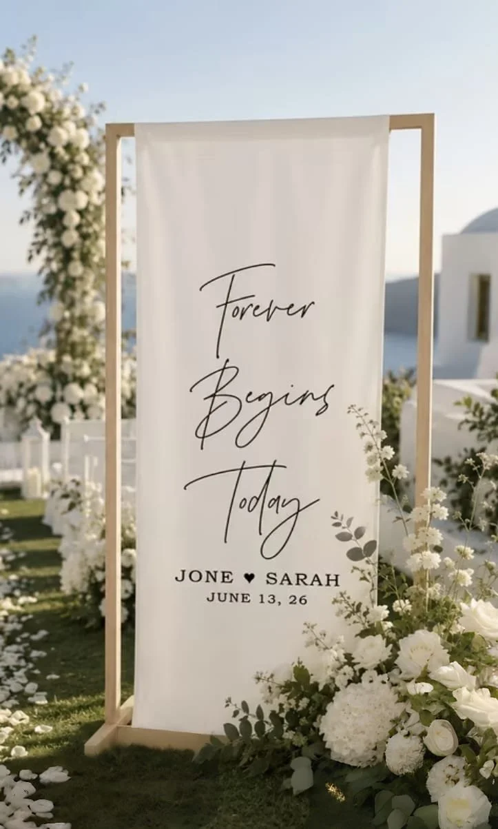 Panneau de mariage blanc avec le texte "Forever Begins Today" et les noms "Jane & Sarah", la date "June 13, 26", entouré de fleurs blanches dans un décor extérieur