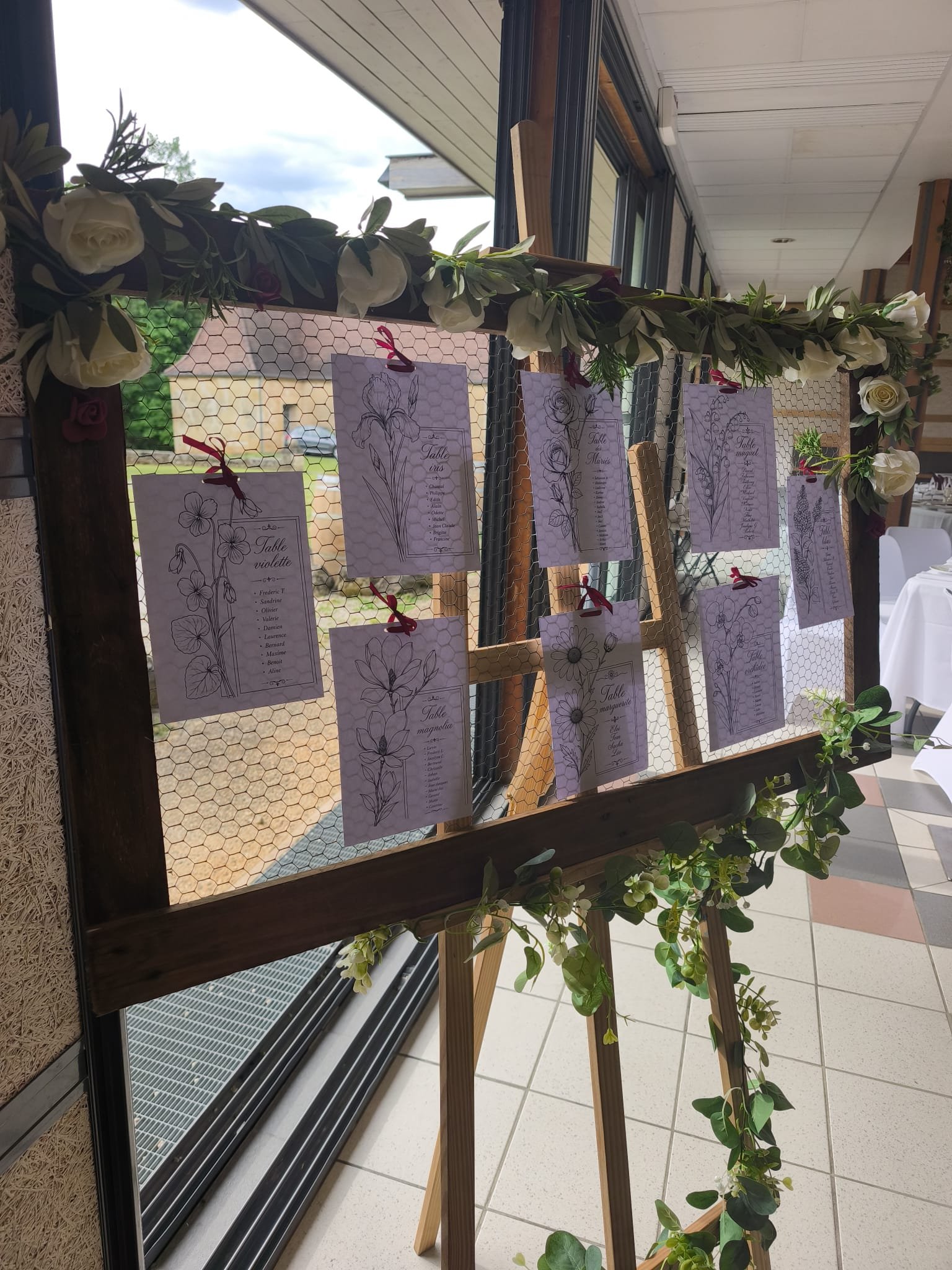 Tableau d'affichage décoré de fleurs blanches et feuillage, avec des feuilles de papier suspendues indiquant des noms de tables pour un mariage ou un événement