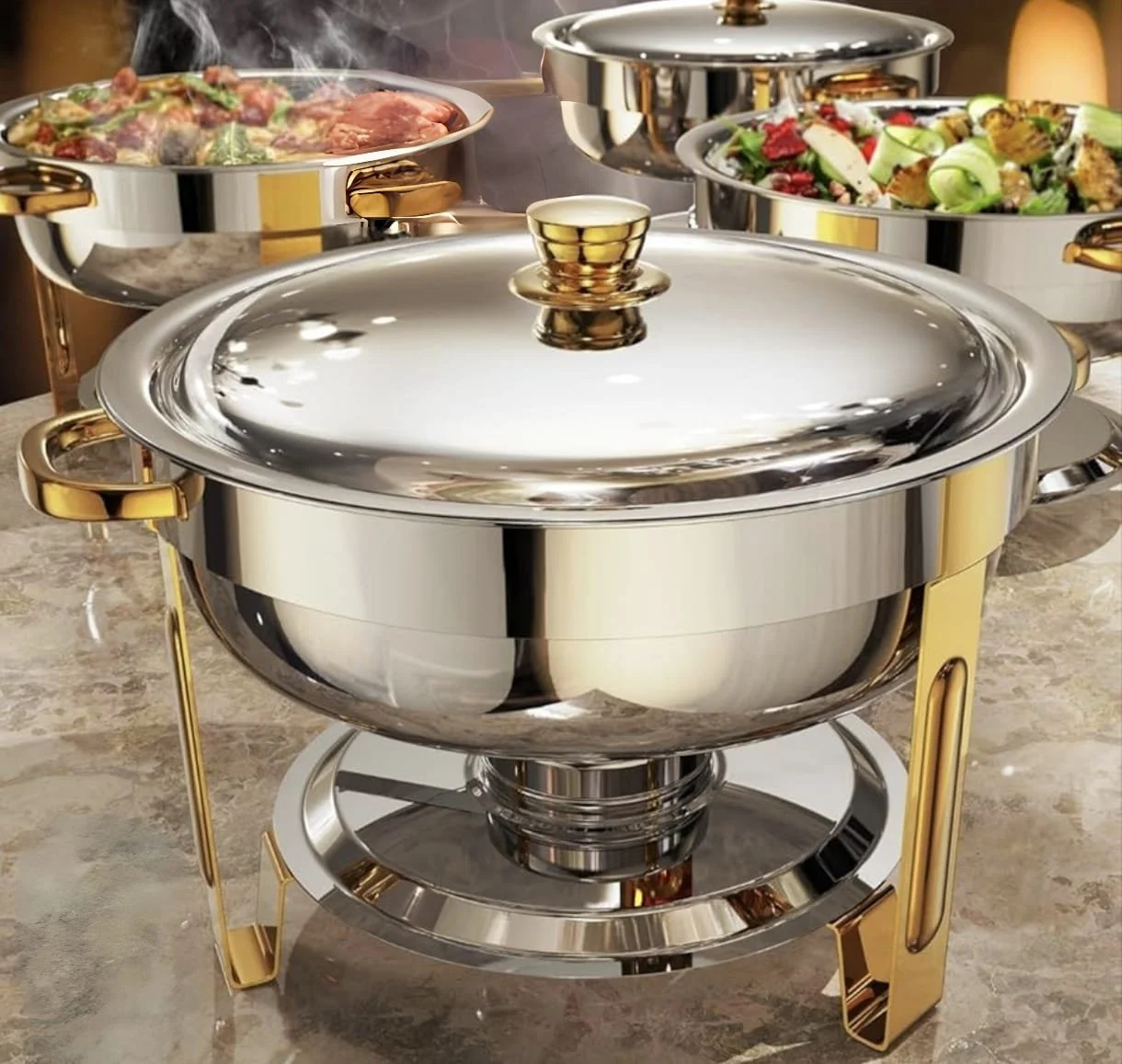 Une table avec des plats chauds en acier inoxydable, y compris un chaudron avec un couvercle, des légumes et de la viande en train de cuire, et une grande salade. La table a une surface en marbre beige.