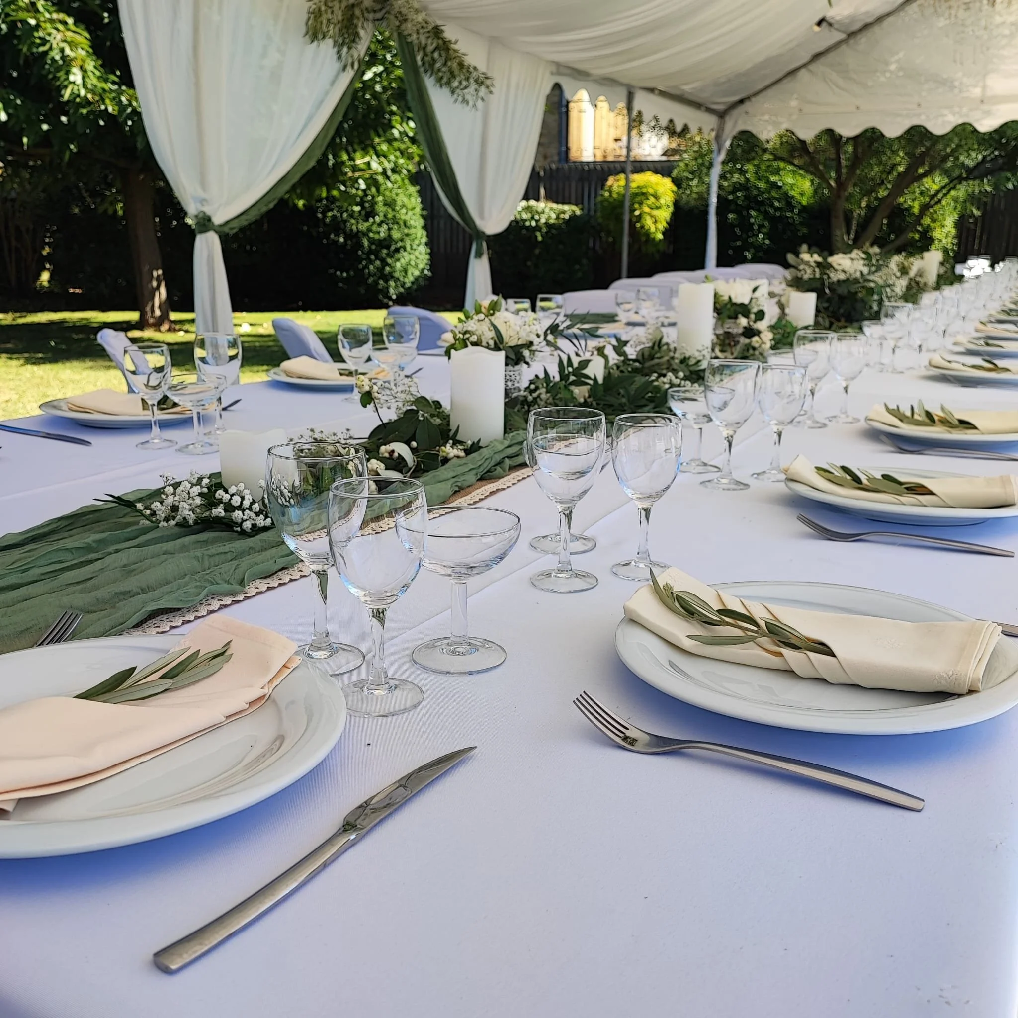 Une table de réception décorée avec des verres à vin, des assiettes, des couverts, des serviettes pliées, une guirlande de feuilles et des bougies blanches, sous une tente blanche en plein air