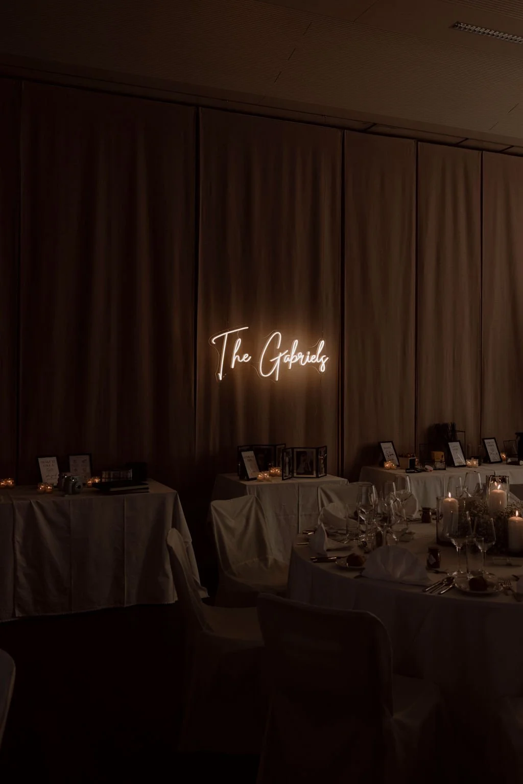 Elegant event space with tables set for a celebration, candles, framed photos, and a neon sign that reads 'The Gabriels' on a large wooden wall.