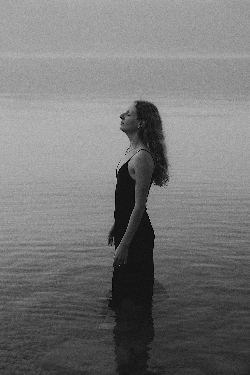 A woman in a sleeveless black dress standing waist-deep in water at the beach with her eyes closed and head tilted slightly upward, black and white photo.