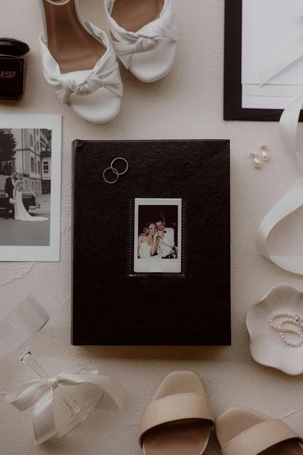 Wedding photo album, a black hardcover book with a Polaroid picture of a bride and groom on the cover, white high-heeled shoes, a glass with a white ribbon, a pair of pearl earrings, a black and white wedding photo, rings, pearl necklace, framed picture, and a bottle of Versace perfume on a white surface.