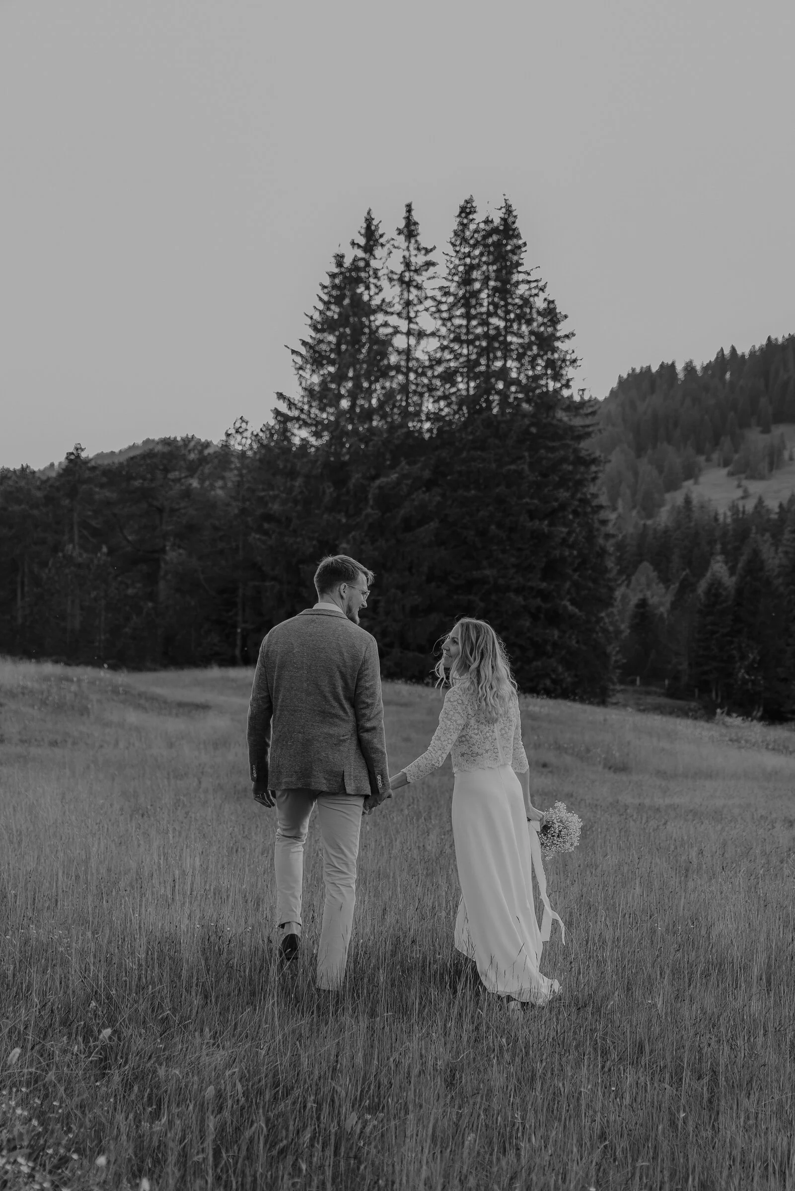 A black and white photo of a man and a woman holding hands in a grassy field with tall trees and mountains in the background. The woman is holding a bouquet of flowers.