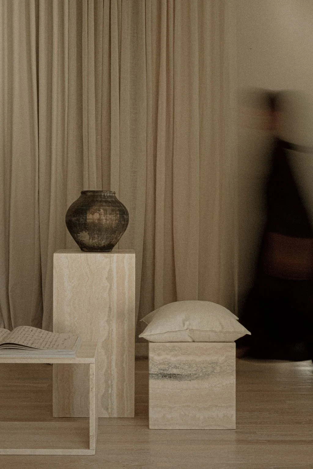 Minimalist interior with beige curtains, a black and brown pottery vase on a marble pedestal, a marble cube with a beige pillow, and part of a white table with an open book.