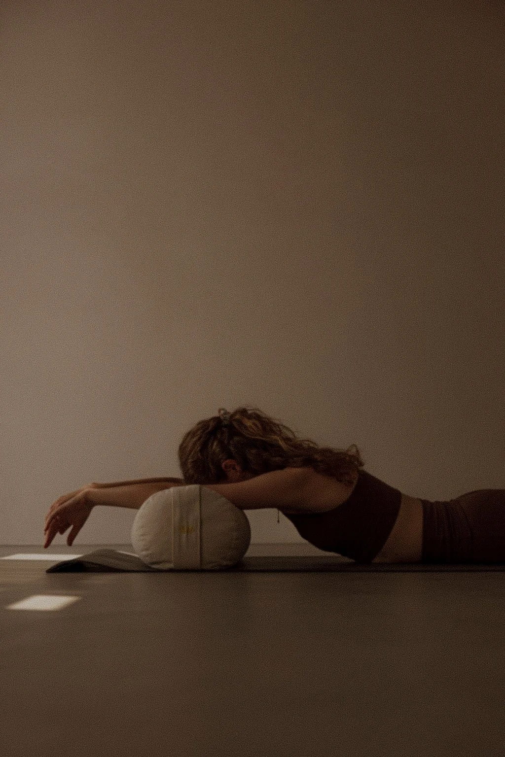 A person practicing yoga, lying face down with their head resting on a cushion, in a dimly lit room.