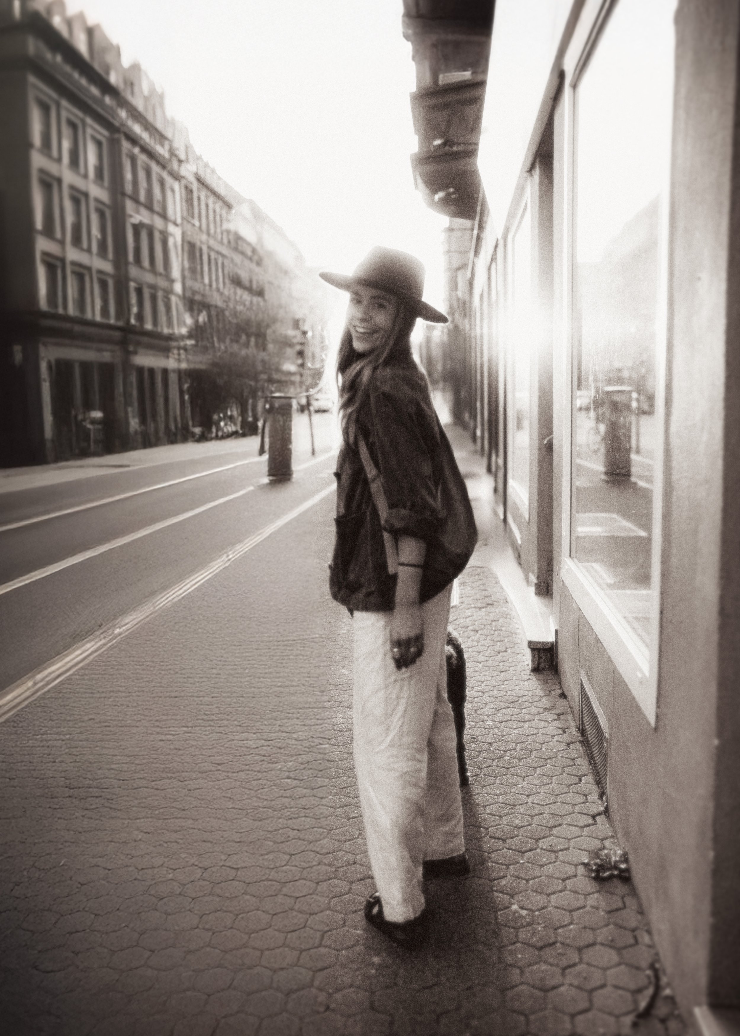 A woman smiling and wearing a large hat, jacket, and pants, walking on a city sidewalk near storefronts and buildings, with a dog nearby.