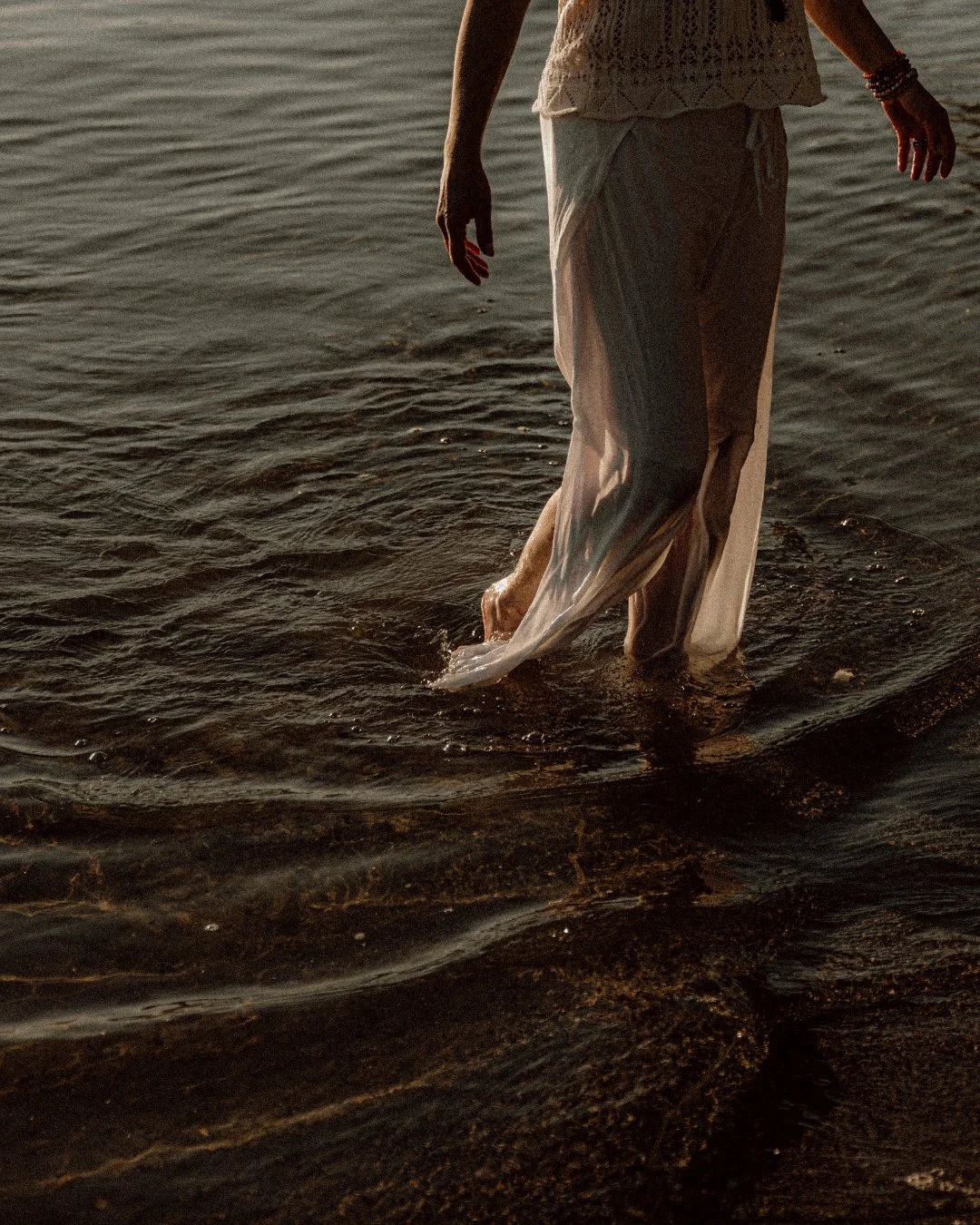 Person walking in water near shore, wearing light-colored loose clothing, with arms slightly extended, during sunset.