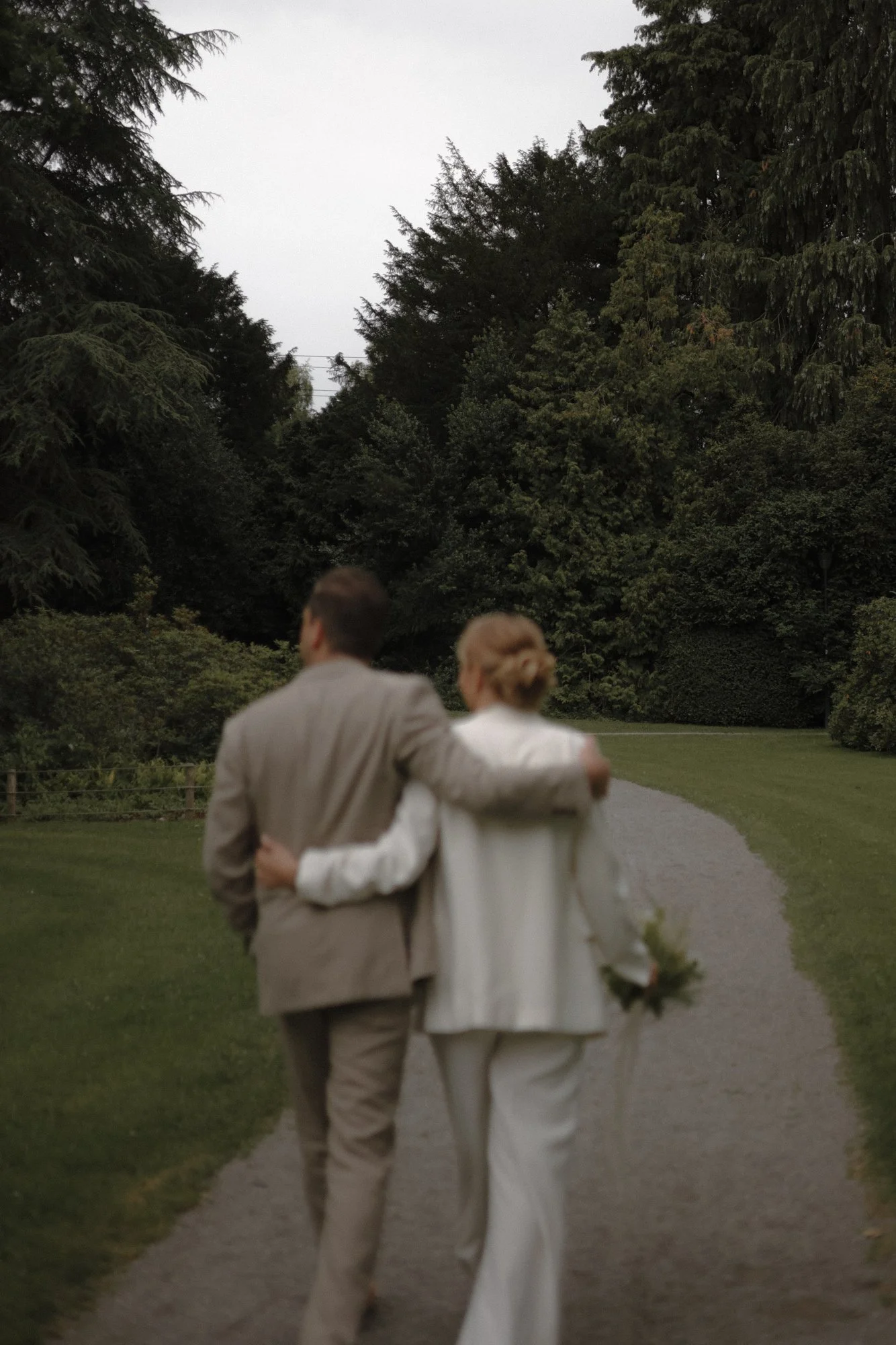 A couple walking arm in arm on a garden path, dressed in formal wedding attire, surrounded by lush green trees and bushes.