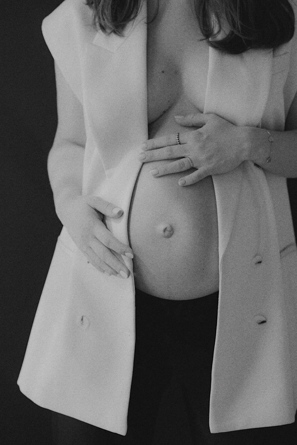 Close-up black and white photograph of a woman's torso, partially covered with an open white blazer, with her left hand touching her belly and her right hand resting on her waist. She has rings on her fingers and a thin bracelet on her left wrist.