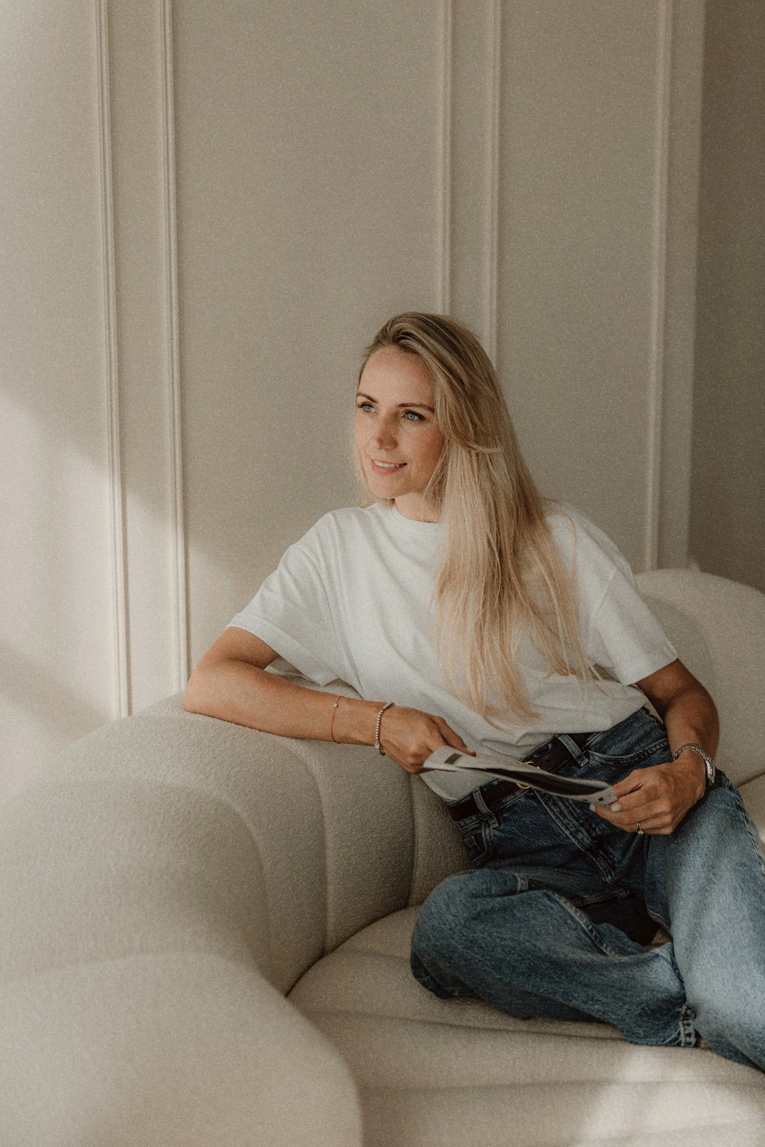 A young woman with long blonde hair, wearing a white t-shirt and jeans, sitting on a beige sofa in a brightly lit room with cream-colored walls, holding a magazine and smiling.