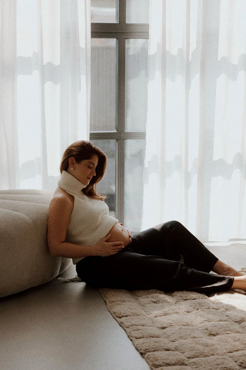 A pregnant woman sitting on the floor near a sofa, gently holding her belly, with a serene expression, in a softly lit room with large windows and sheer curtains.