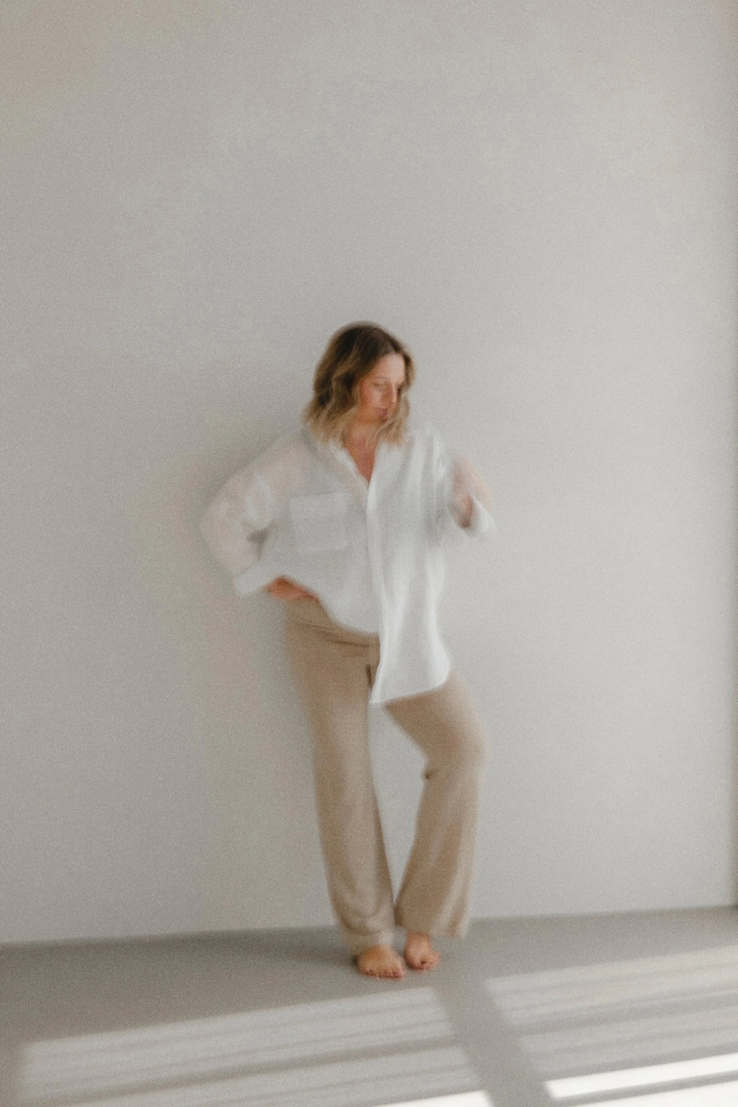 A woman standing in front of a plain white wall, wearing a white blouse and beige pants, with her left hand on her hip and her right hand adjusting her hair.