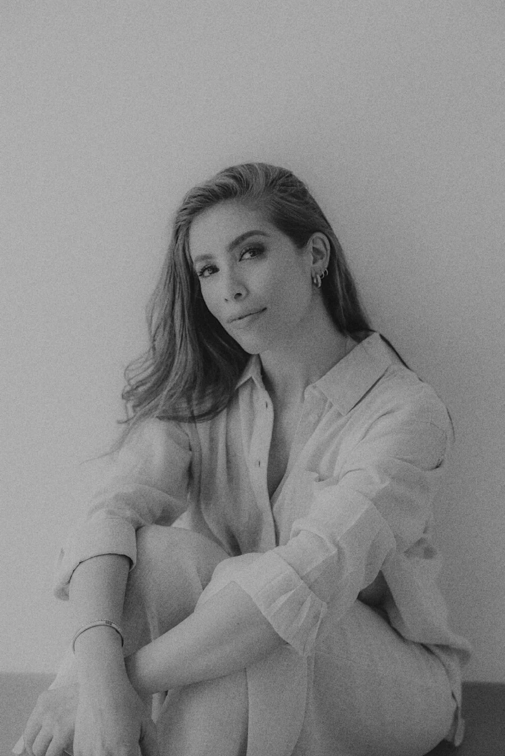 A black and white photo of a woman sitting against a plain wall, wearing a button-up shirt with sleeves rolled up. She has long hair, earrings, and a bracelet, and looks at the camera with a slight smile.