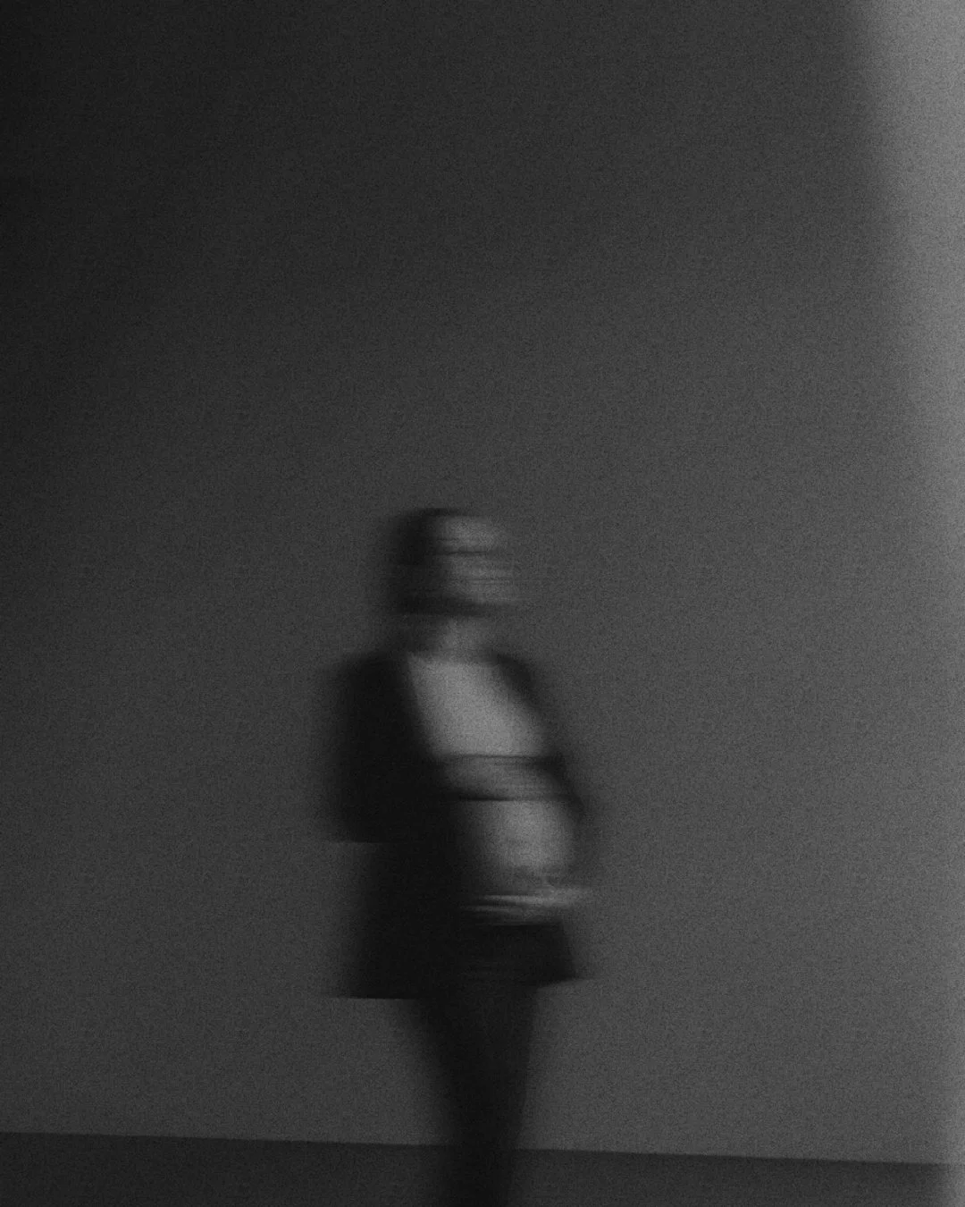 A blurry, black-and-white photo of a person standing against a wall with their arms crossed, facing slightly to the right.