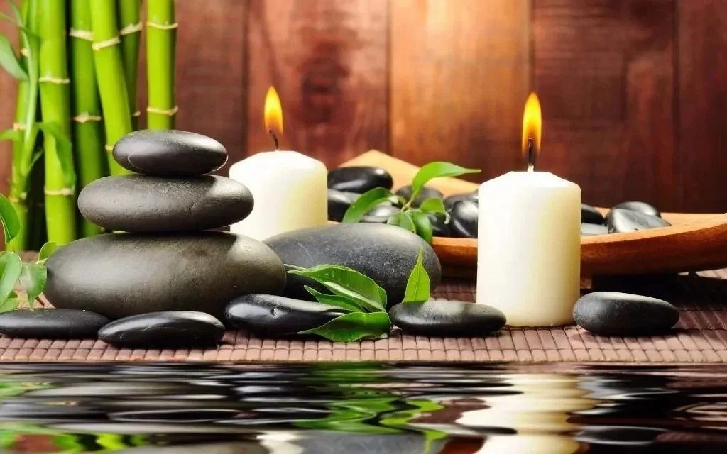 Stacked black massage stones, white candles with flames, green leaves, bamboo, and water on a bamboo mat, creating a spa or relaxation setting.