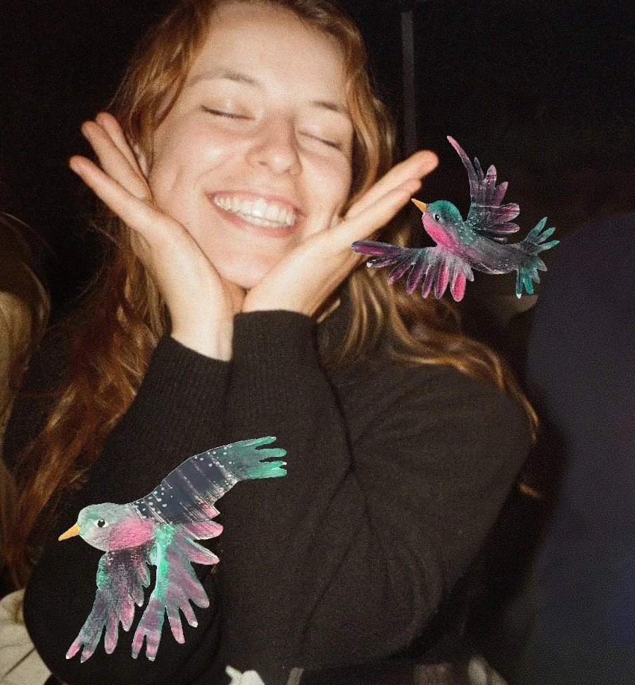Young woman smiling with eyes closed, holding her face with both hands, with two colorful illustrated hummingbirds near her face and arm.