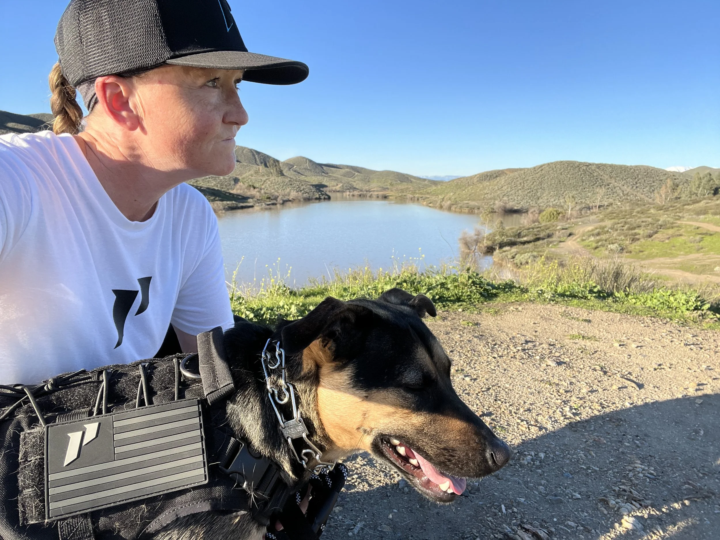 A woman wearing a baseball cap and white shirt sitting outdoors with a black and tan dog, with a lake and rolling hills in the background on a clear day.